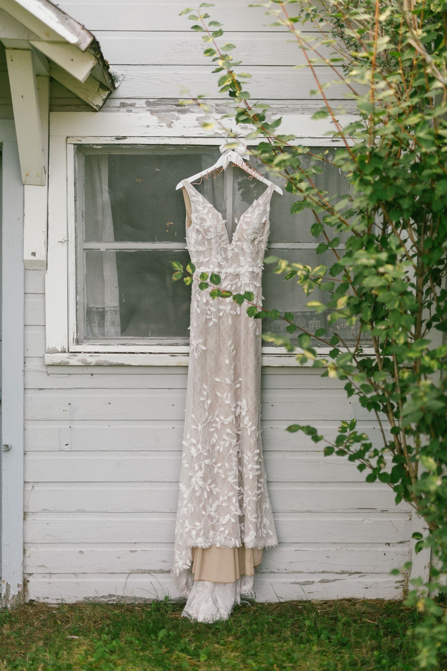 A delicate lace wedding dress hangs on a white hanger against a rustic white wooden wall with green foliage.
