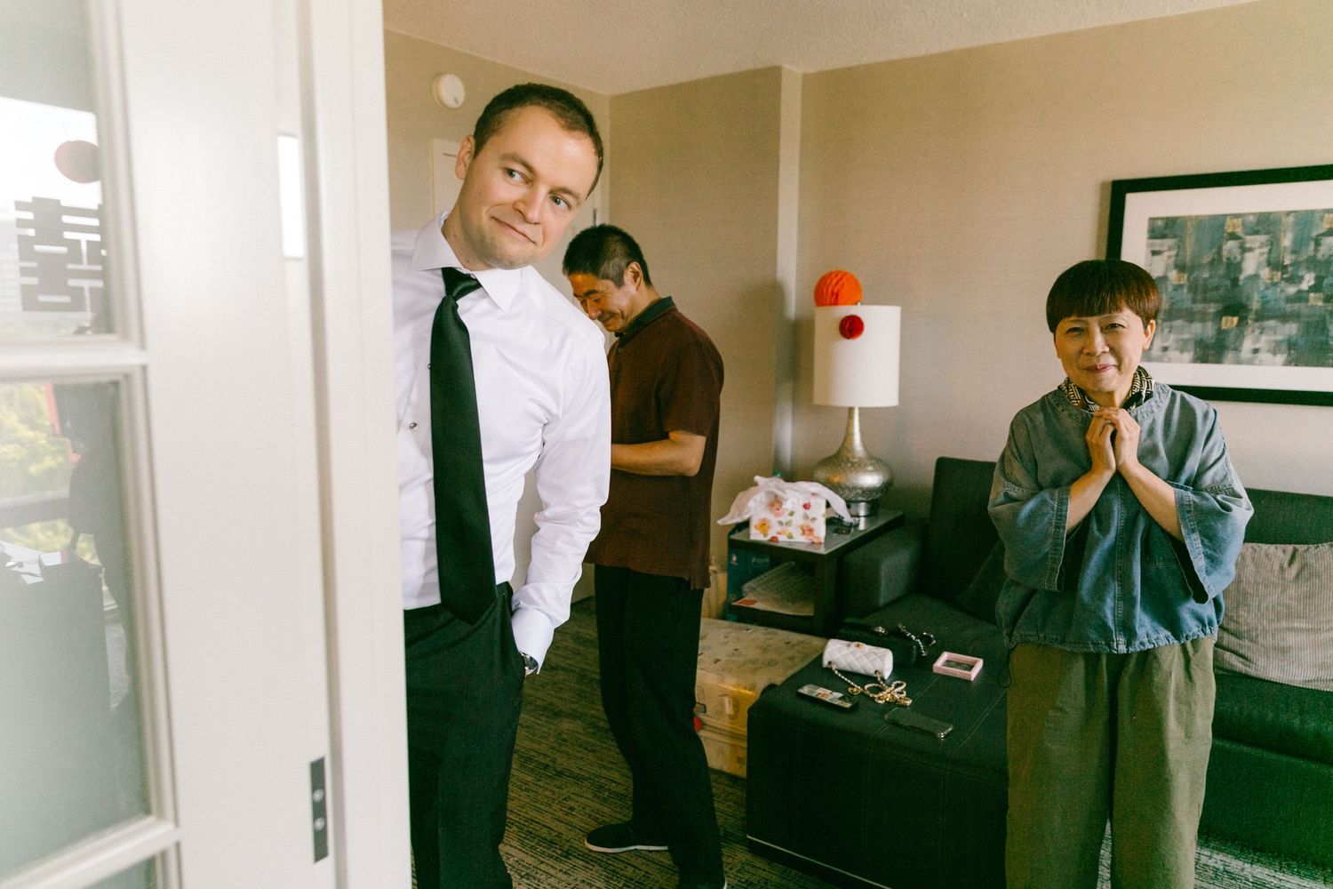 Three men in a hotel room, one in a white shirt and tie, another in casual wear with hands clasped, preparing for an event.