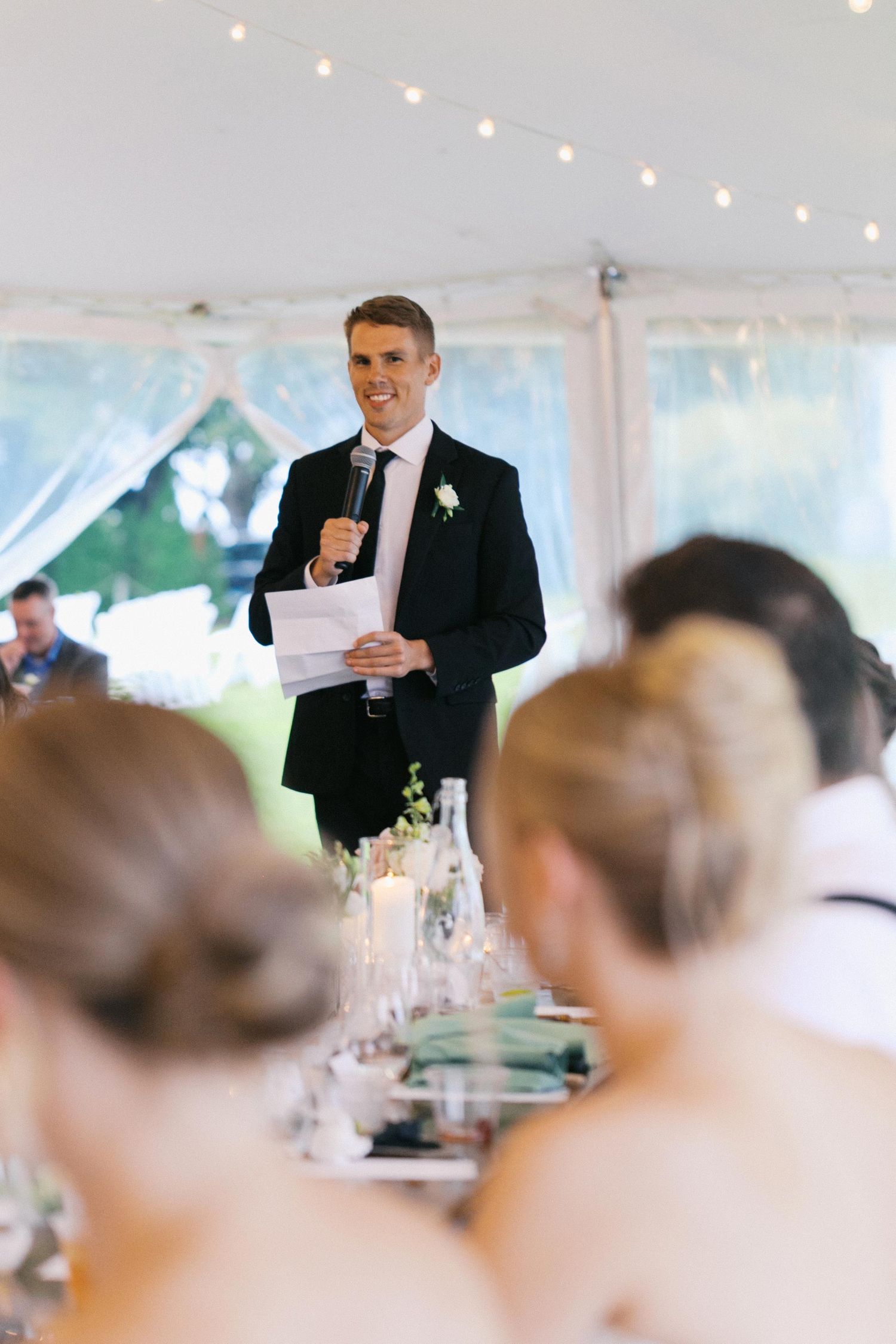 A wedding reception speech being delivered in a white tent with guests seated at tables.