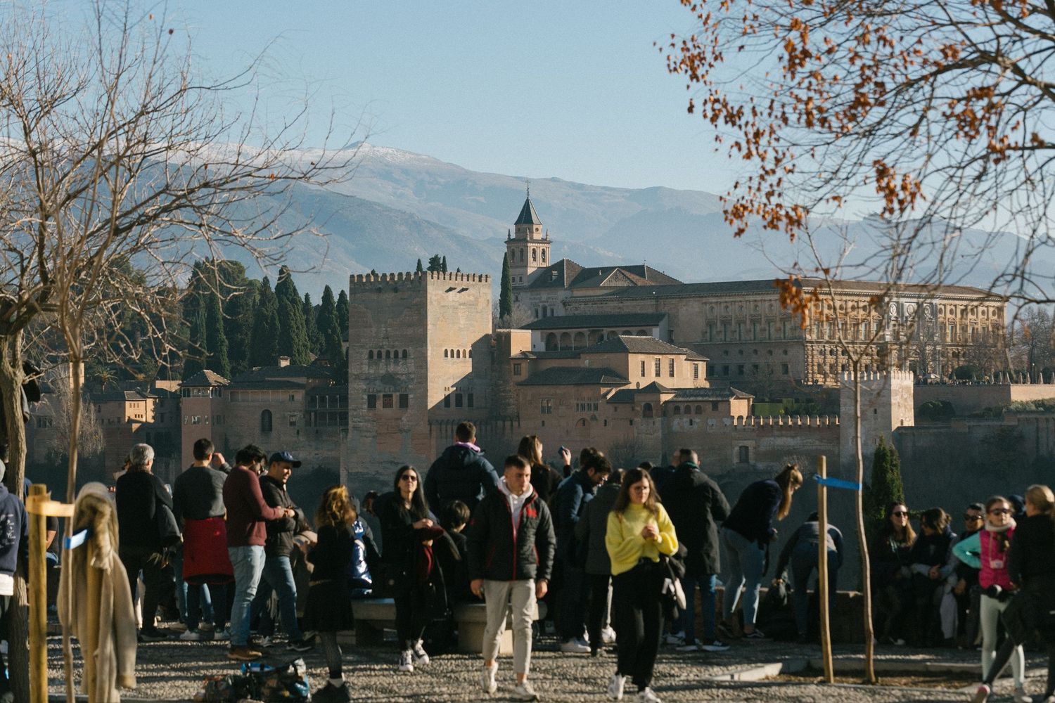Tourists gather at a viewpoint overlooking the historic Alhambra palace fortress in Granada, Spain with mountains in background.