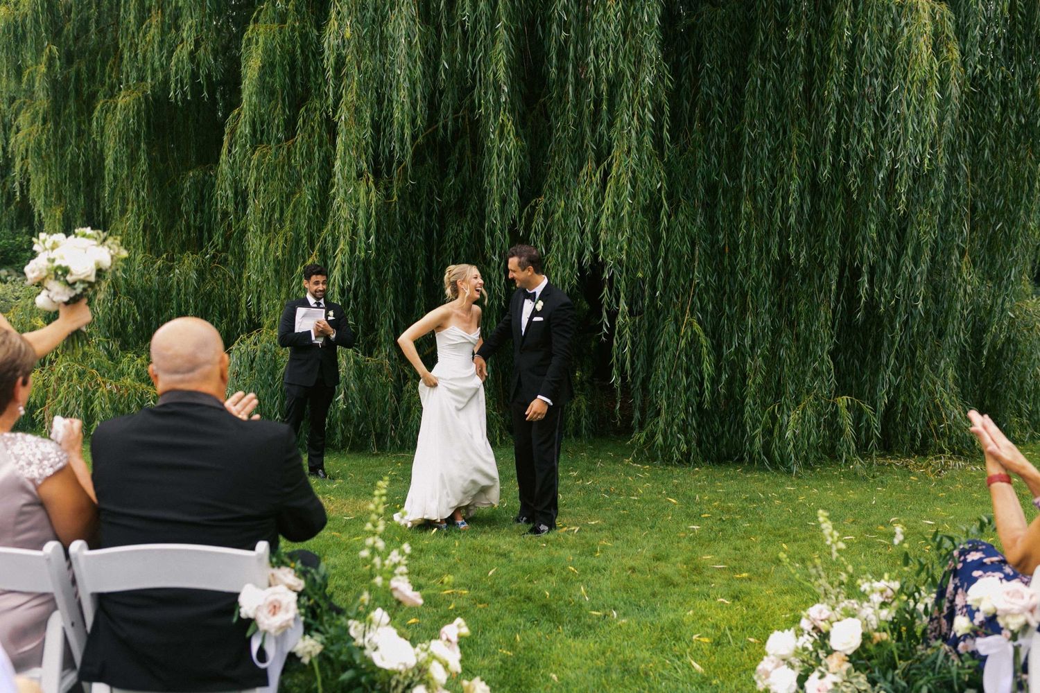 A panoramic view of a wedding celebration on a lawn with weeping willow trees creating a romantic backdrop.