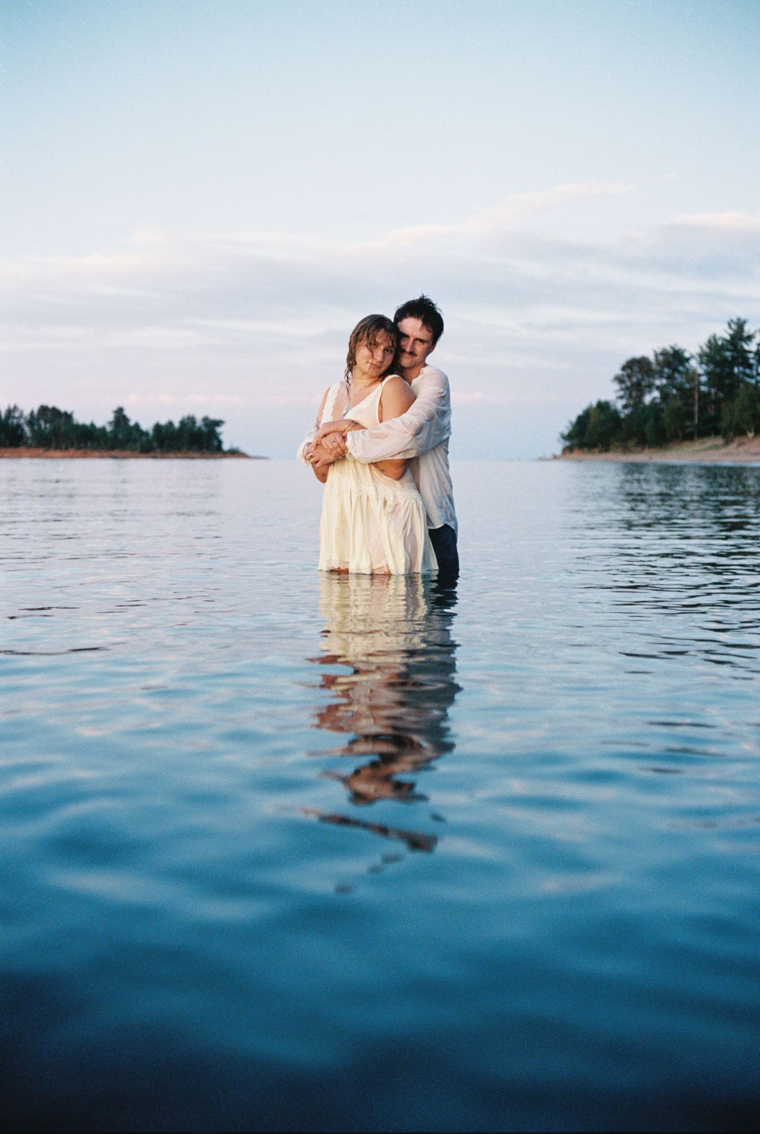 Couple embracing romantically while standing in calm shallow water near a serene shoreline at dusk.