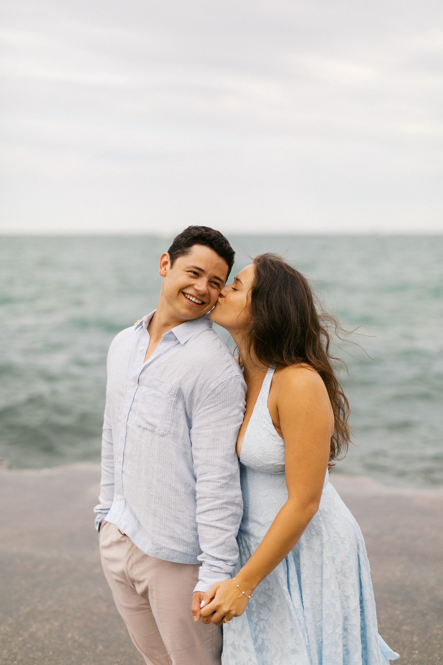 Couple holding hands by the ocean, woman in blue dress kissing mans cheek as he smiles warmly.
