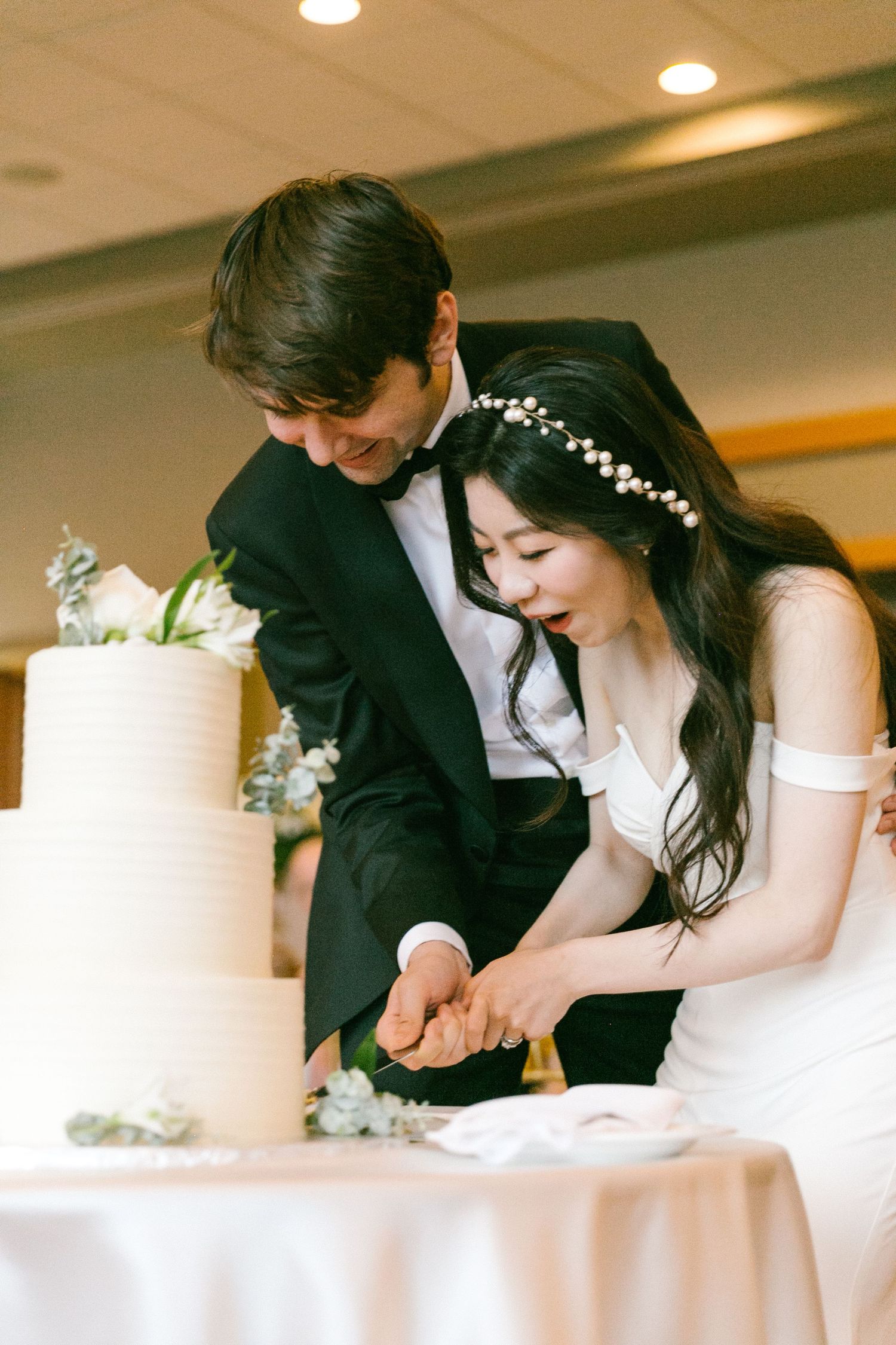 Bride and groom cut elegant three-tier white wedding cake together at their reception celebration.