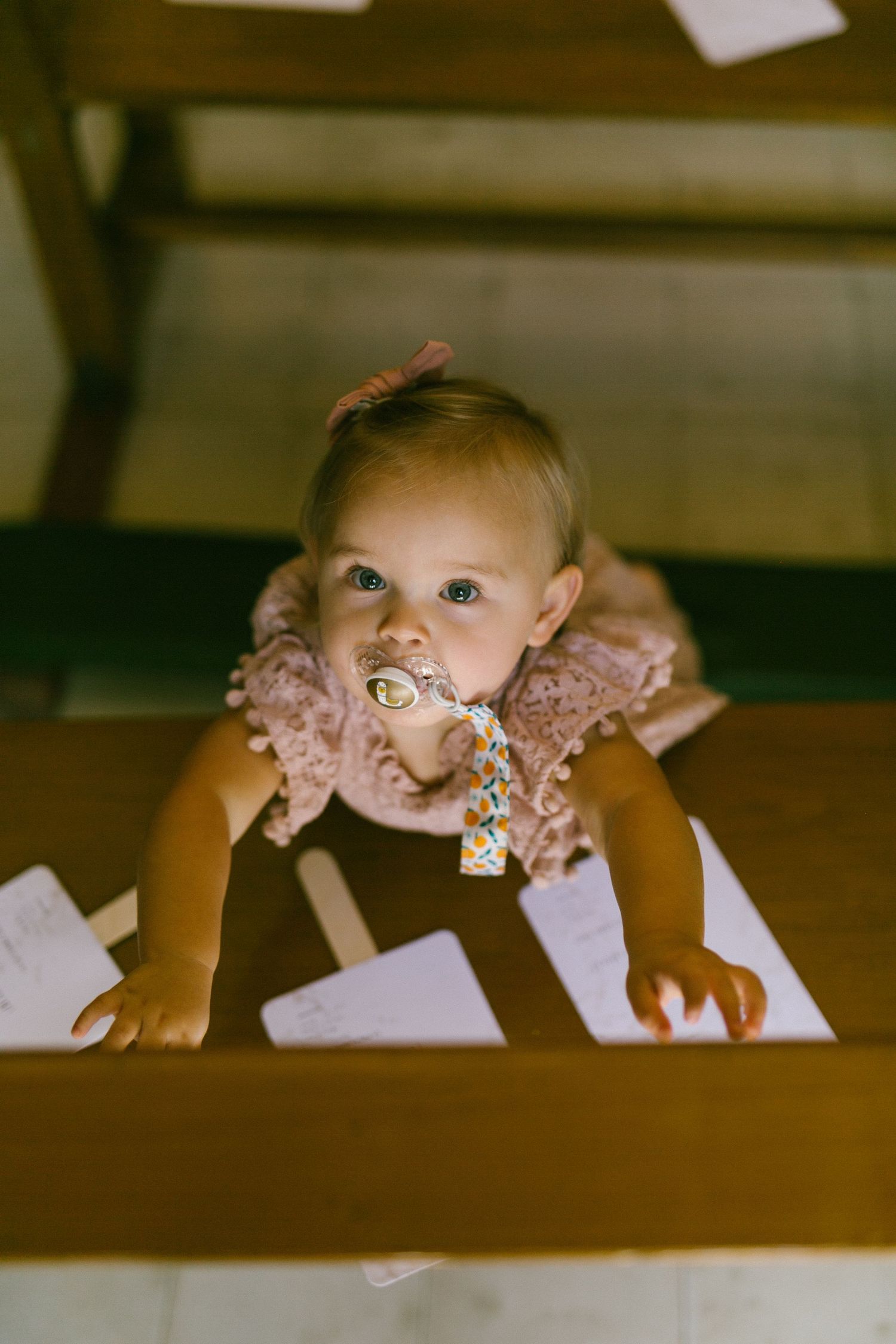 Baby in a pink lace dress crawls over scattered papers on the floor in vintage-toned photograph.