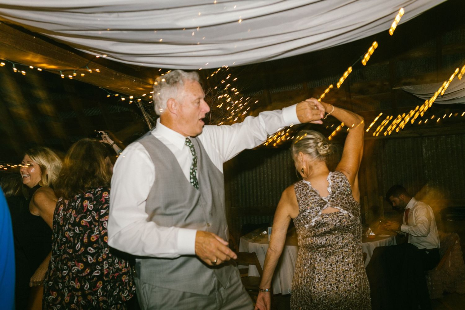 Two people dance together at an indoor celebration under white fabric draping and string lights.