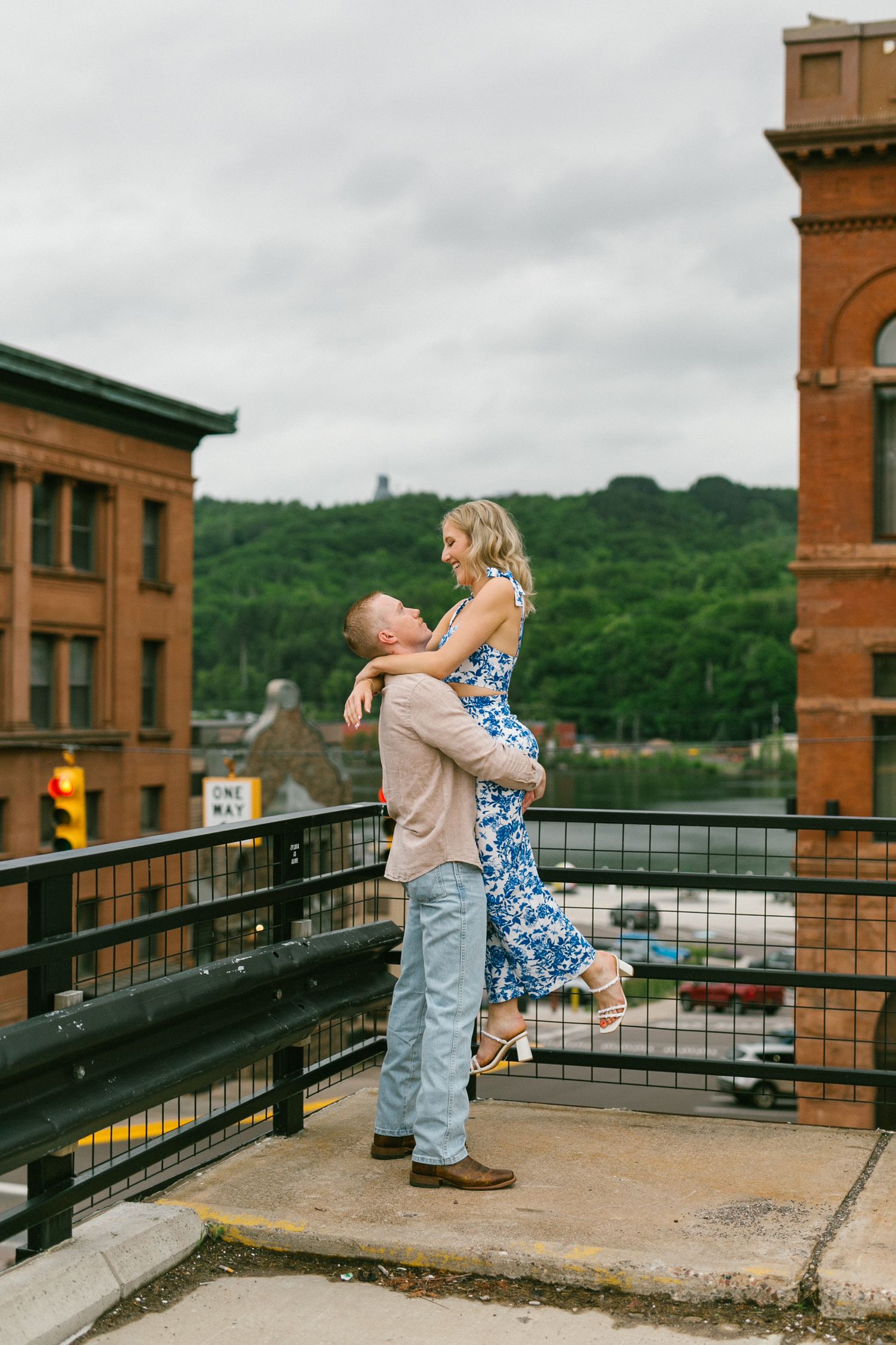 A romantic moment between a couple embracing on an elevated walkway with brick buildings and green hills in background.