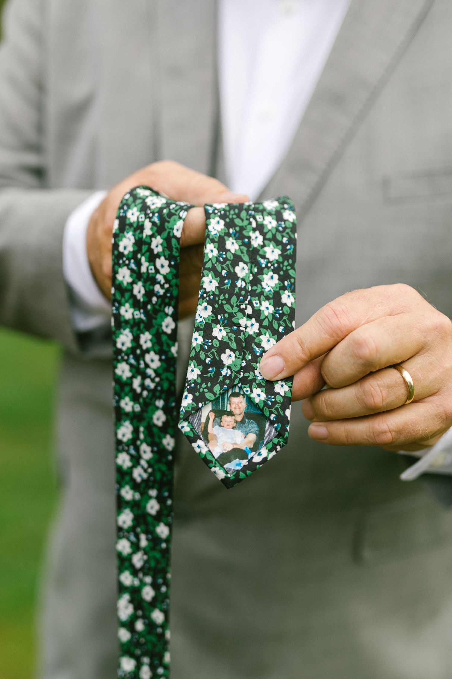 Father of the bride holding a green and white floral patterned customized tie against a gray suit background.