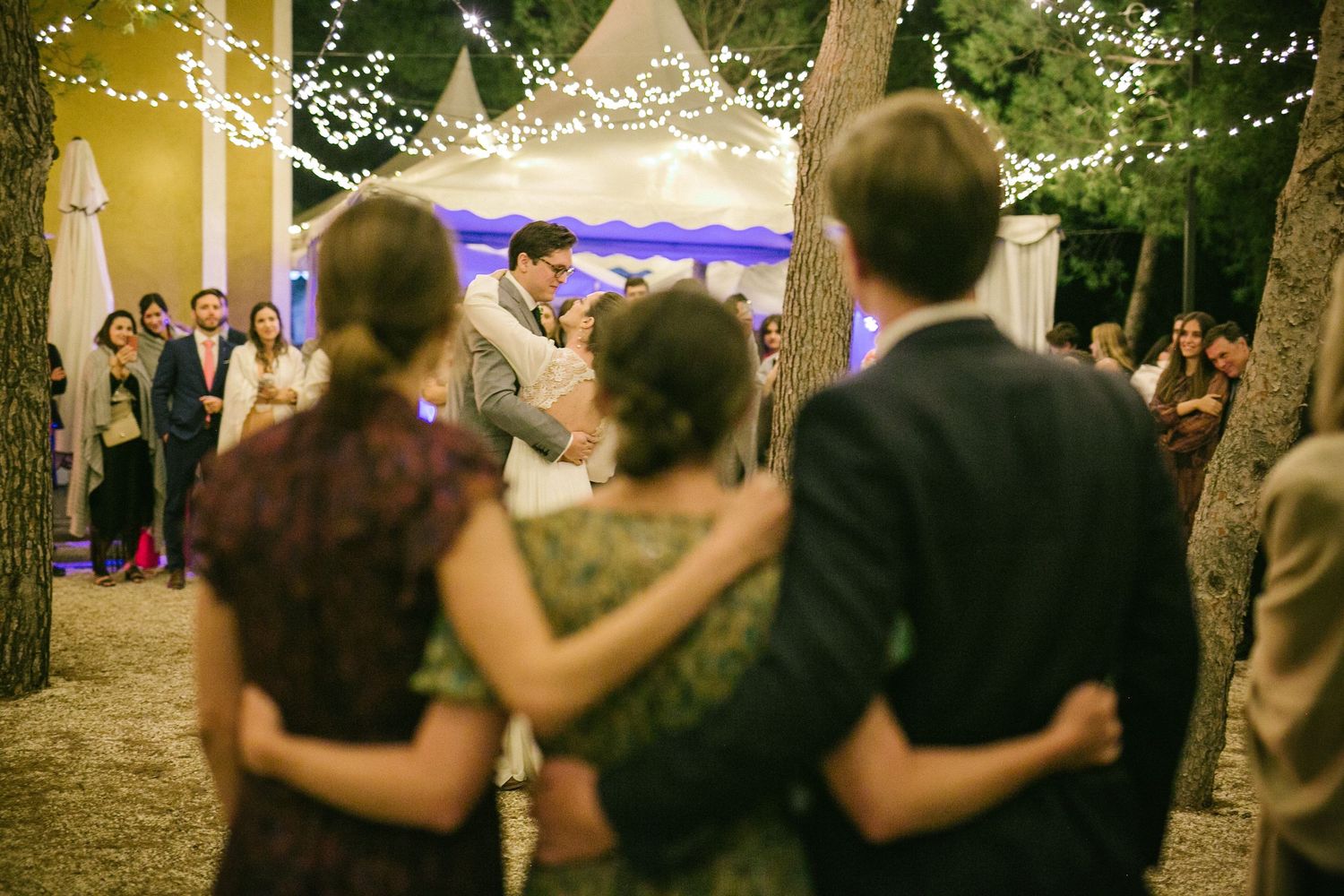 Wedding guests observe and embrace while watching a dance under string lights at an outdoor evening reception.