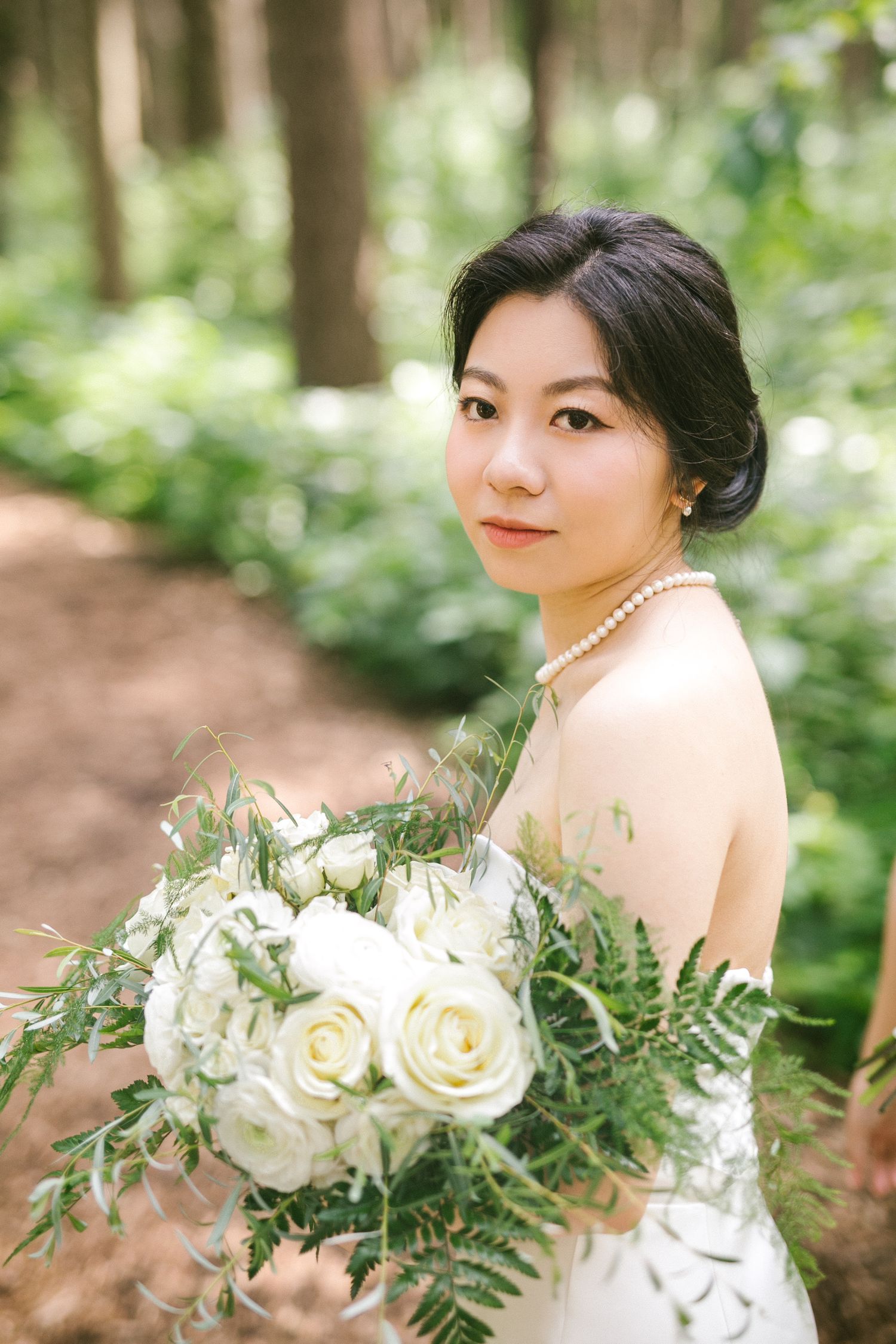 Bride in white strapless gown holding white rose bouquet with ferns in a lush green forest setting.