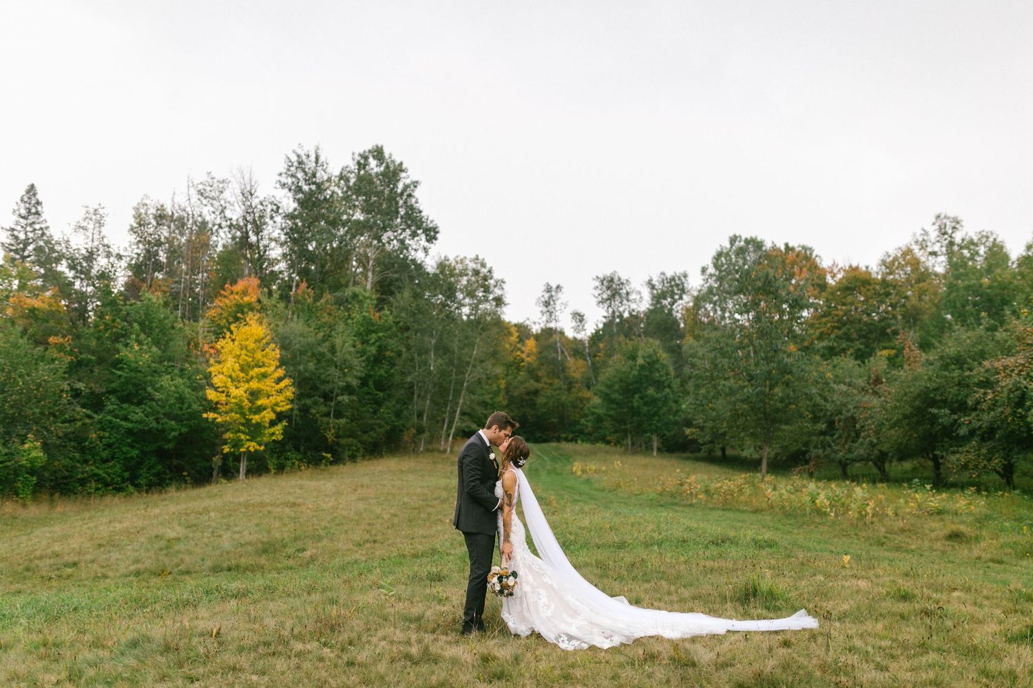A couple stands together in a scenic fall meadow with trees and rolling hills in the background.