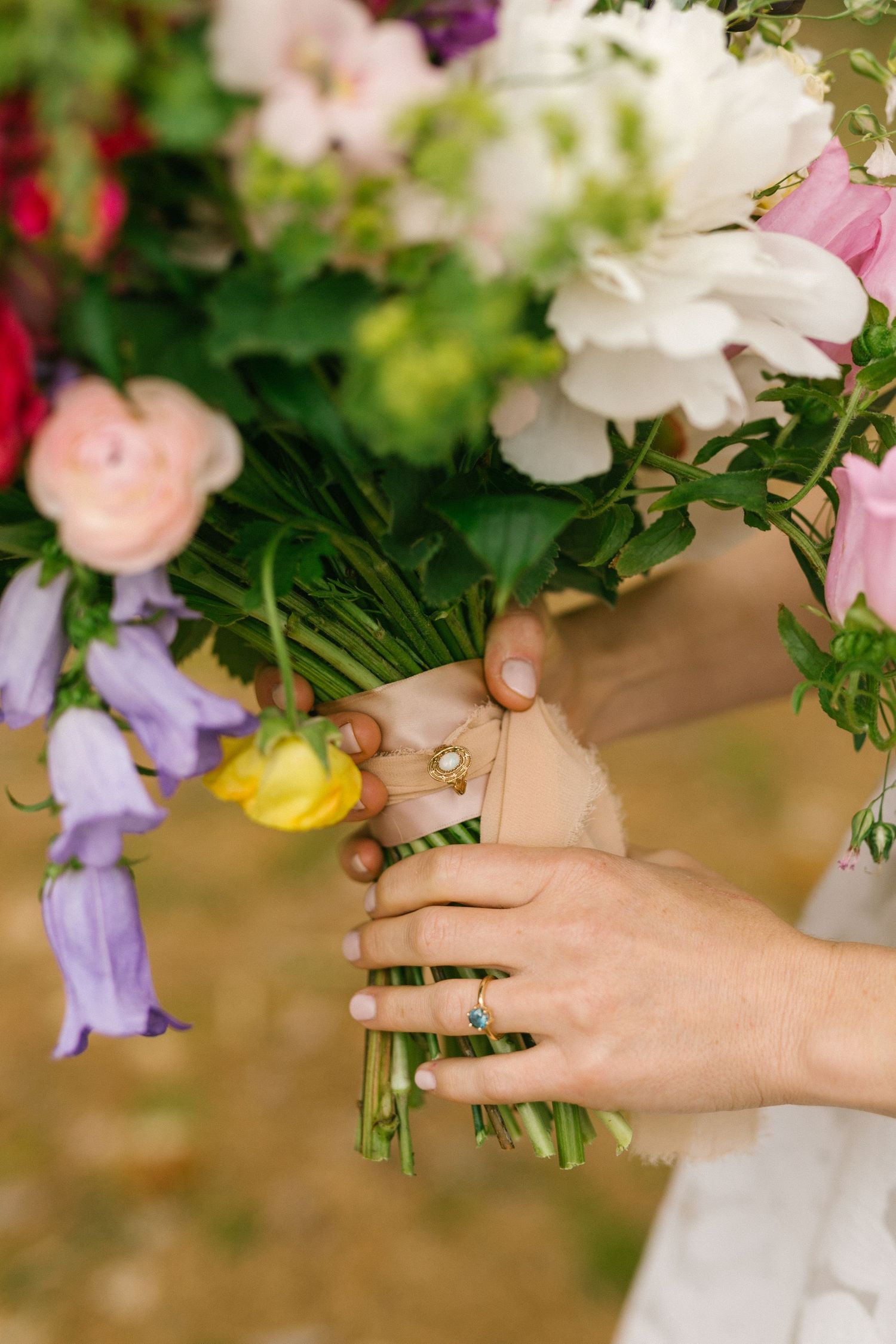 Hands holding a vibrant bouquet of mixed flowers including white, purple, pink and yellow blooms against a blurred background.