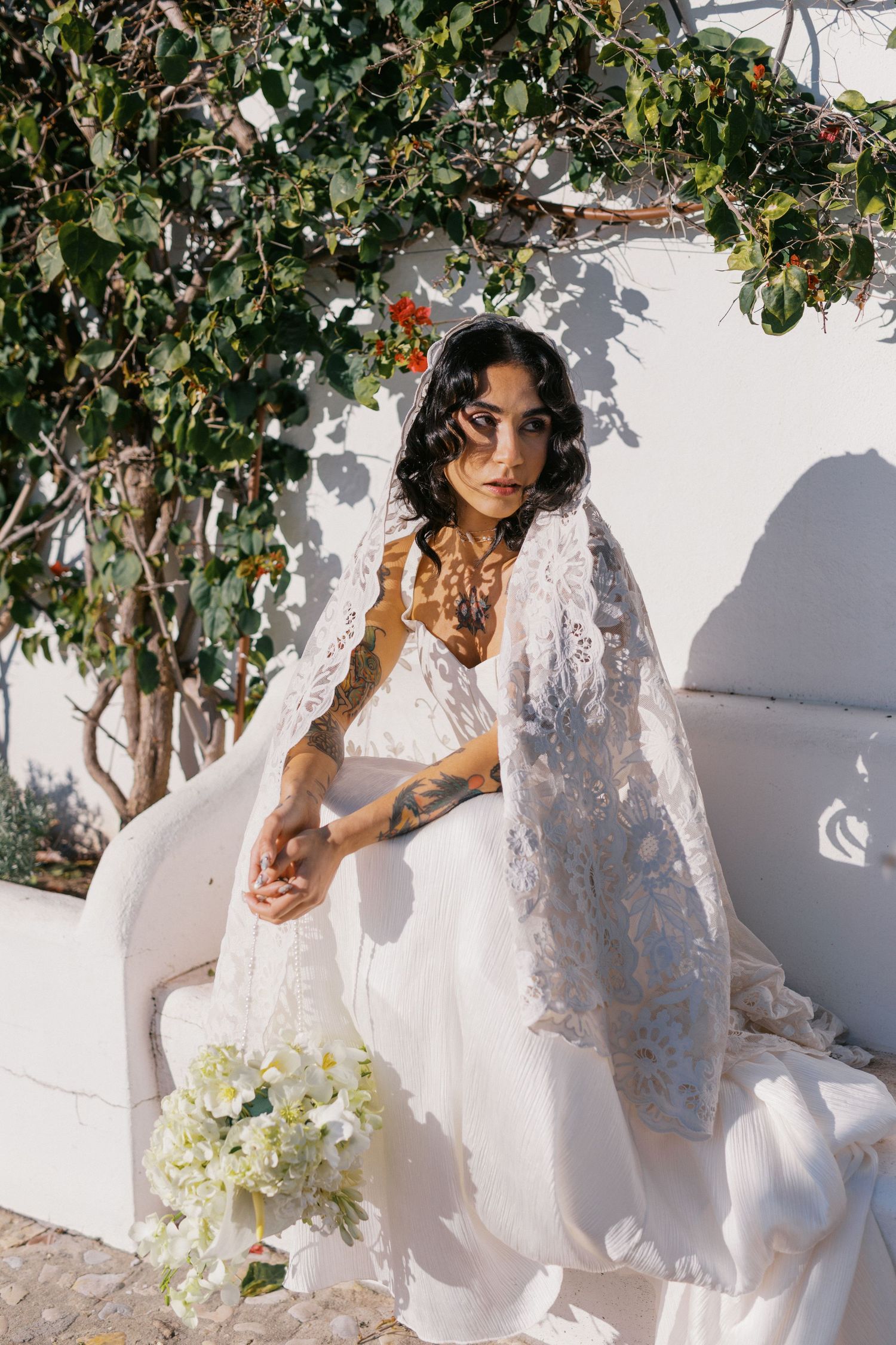Tattooed bride in white gown and lace veil seated outdoors holding white flower bouquet near greenery.
