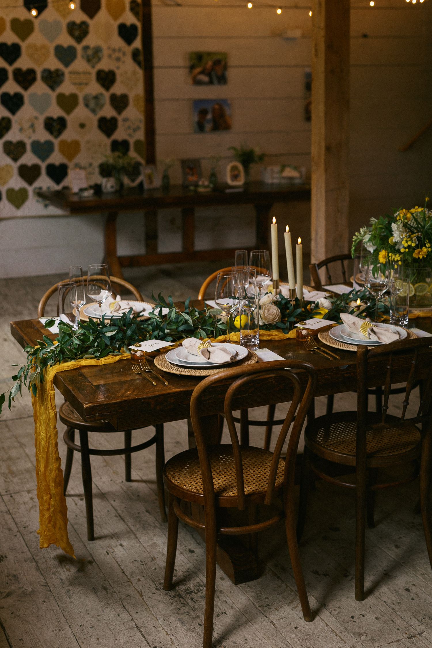 Rustic wedding reception table with mustard runner, greenery garland, white candles, and elegant place settings in a barn venue.