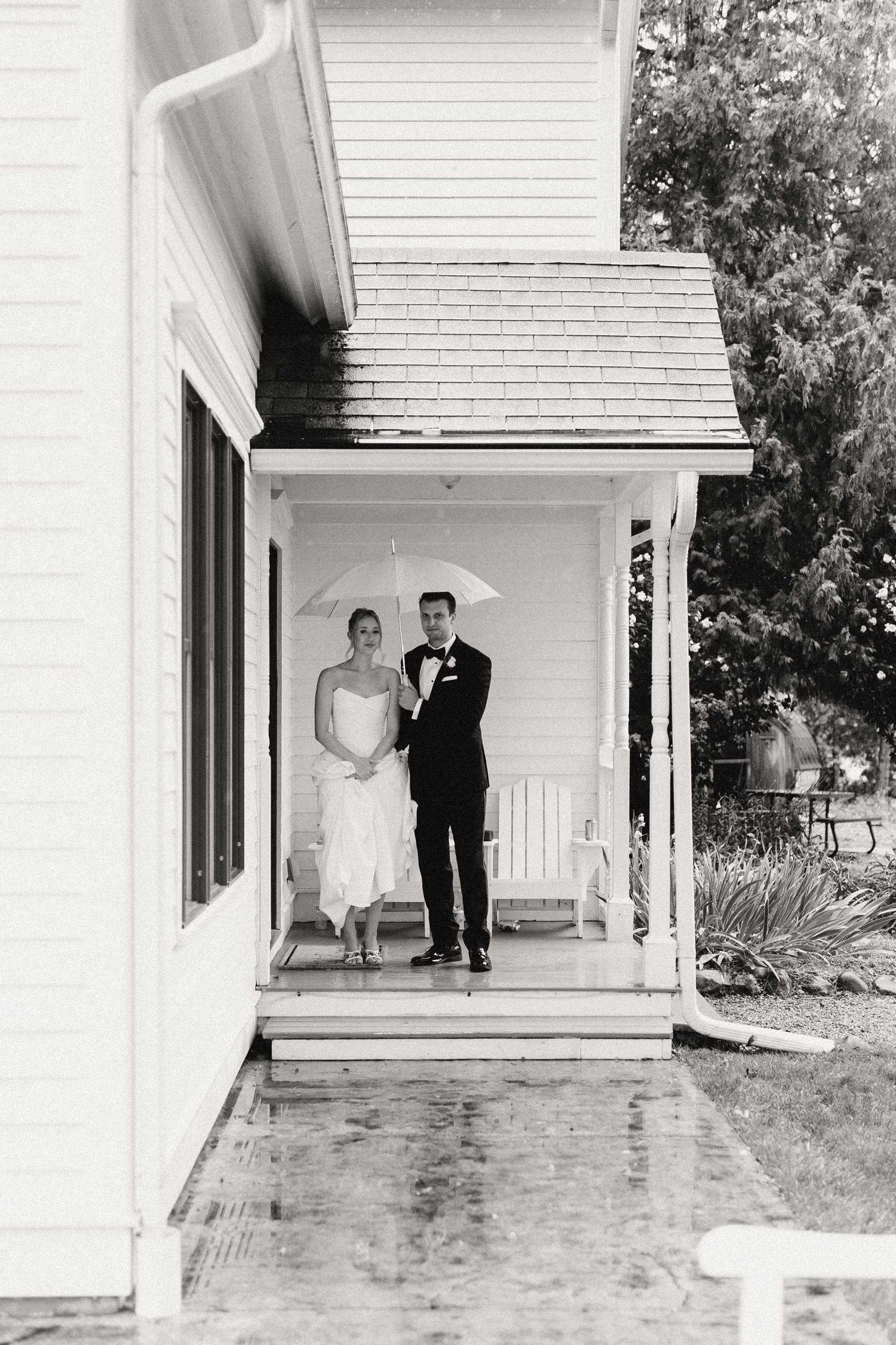 A bride and groom stand on a white porch of a traditional house while waiting to walk through the rain.