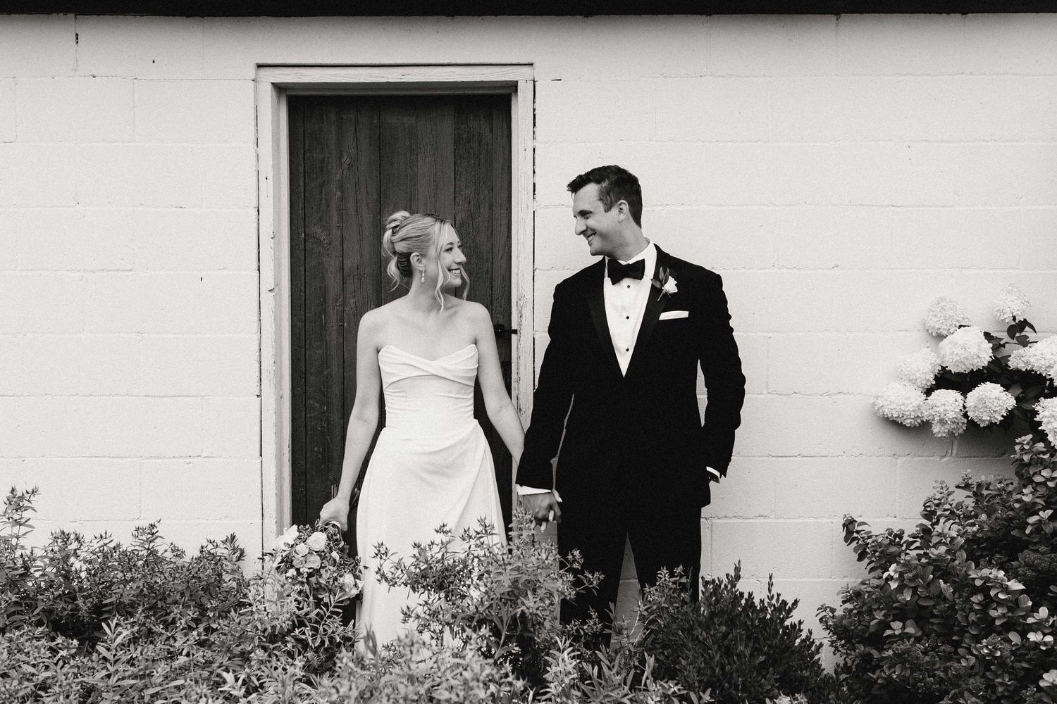 A black and white wedding photo showing couple walking and posing against a wall with flowers.