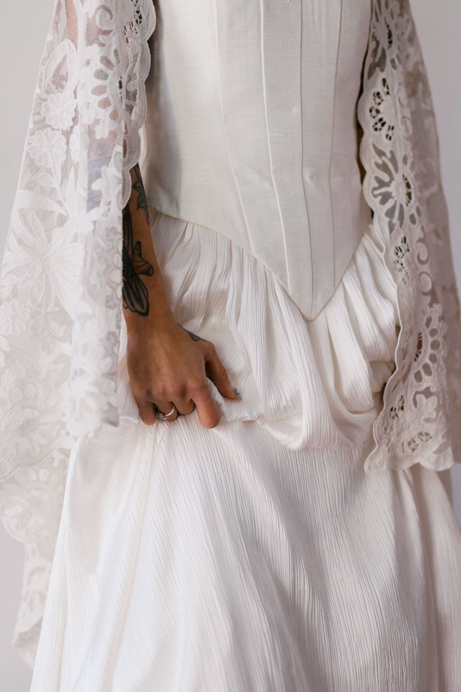 Close-up of bride in white corset wedding dress with intricate lace veil, showing tattooed hand holding skirt.