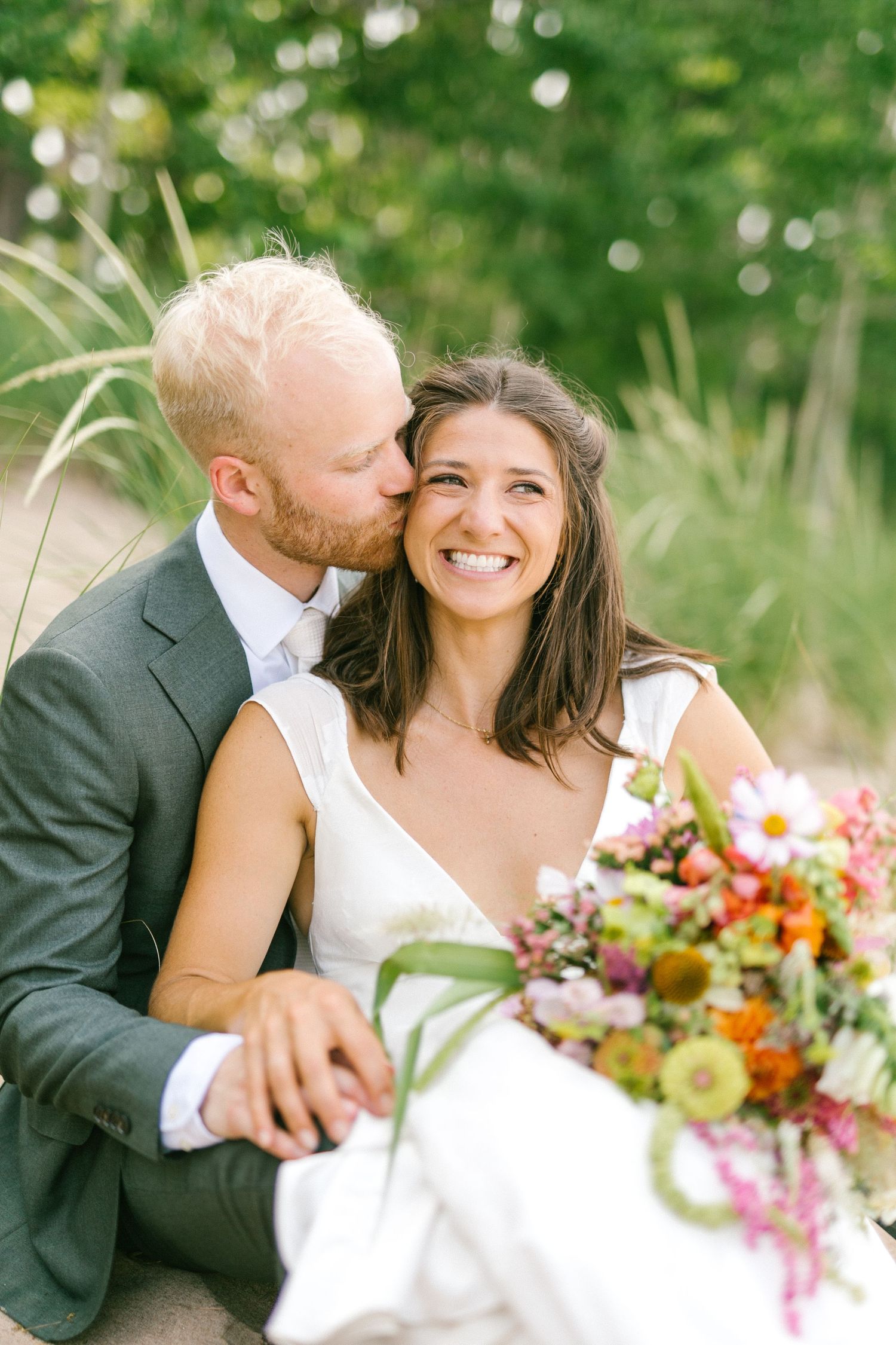 Joyful wedding couple shares tender moments outdoors surrounded by summer greenery and holding vibrant flowers.