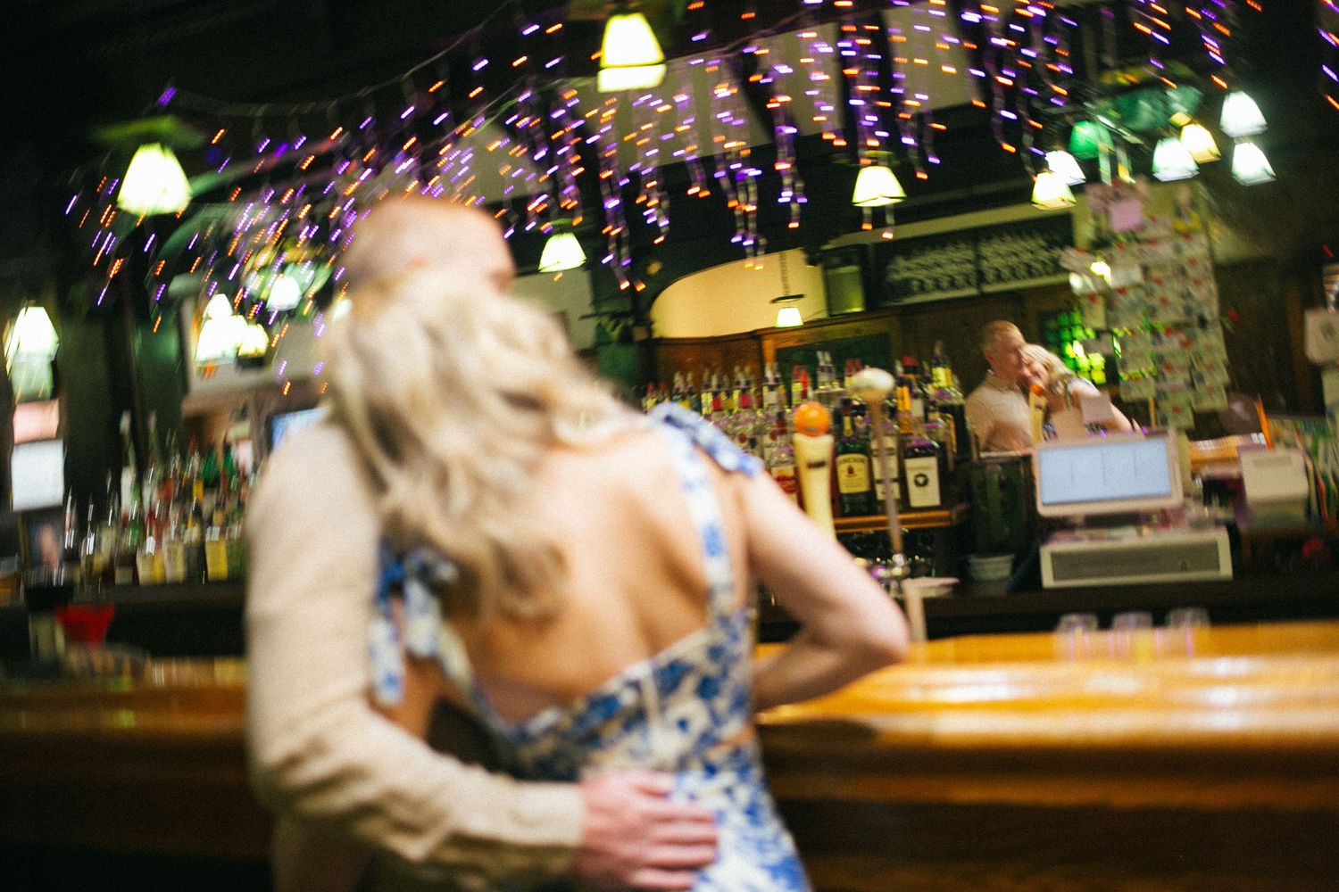 Blurred motion shot inside a dimly lit bar with festive string lights and glowing lamps overhead.