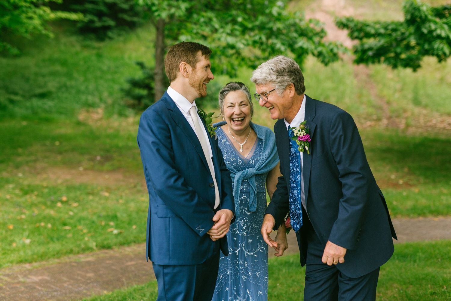 Three people share a joyful candid moment together while dressed formally in navy suits and a blue dress outdoors.