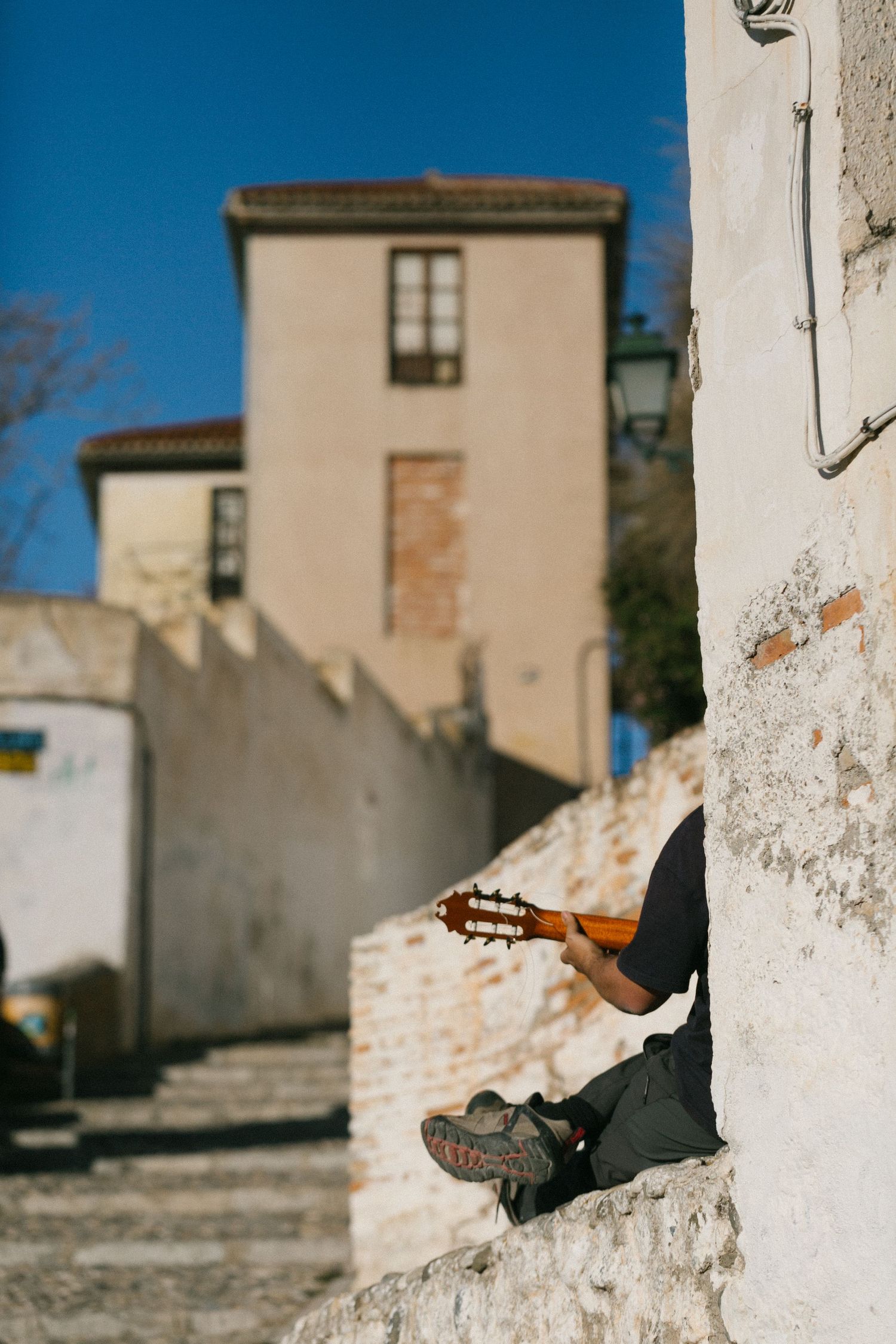 A musician plays guitar while sitting on white stone steps against a beige building with a blue sky backdrop.