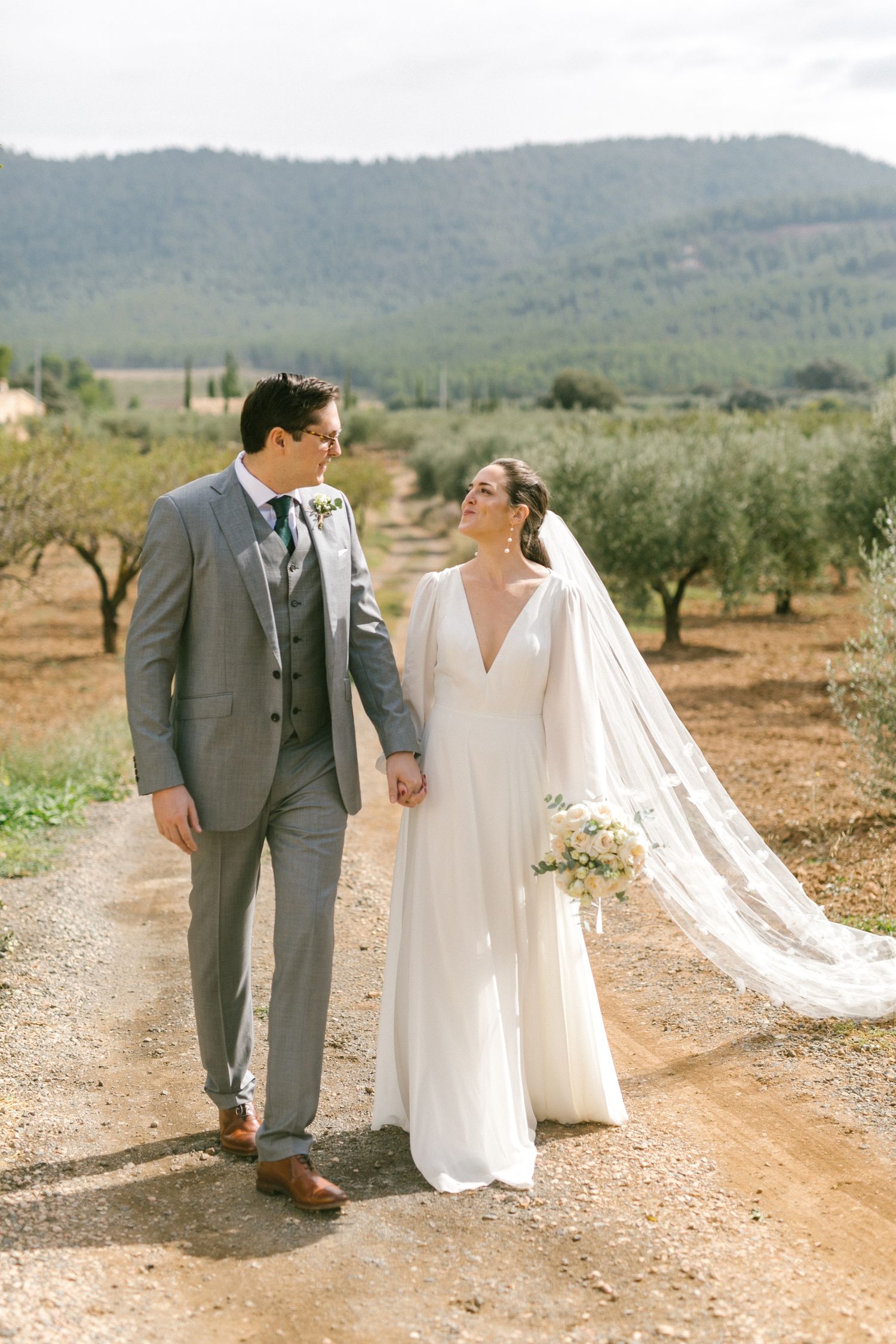 Newlywed couple walking hand in hand on a dirt path through an olive grove with mountains in the background at Spain destination wedding.