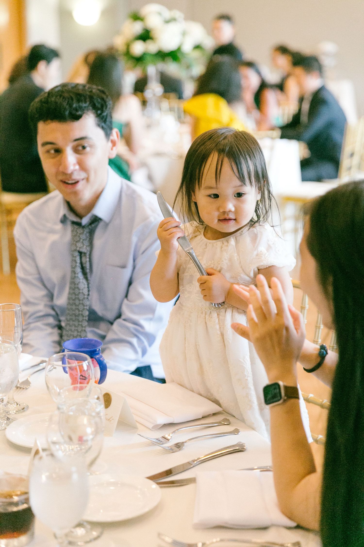 Adorable toddler girl in white dress holds cutlery at wedding reception dinner table with guests.