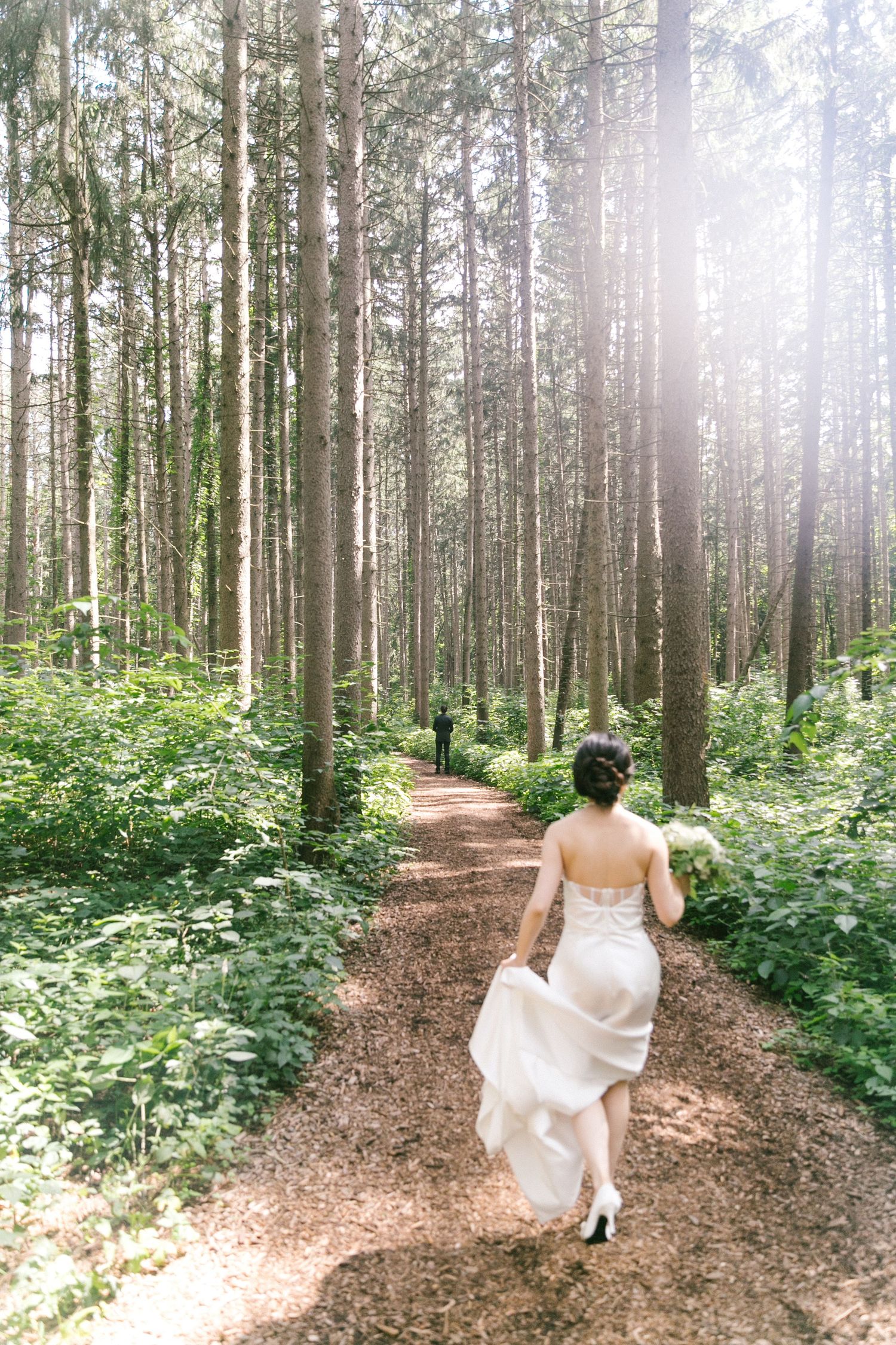Bride in white gown walking along a sunlit forest path toward her groom.