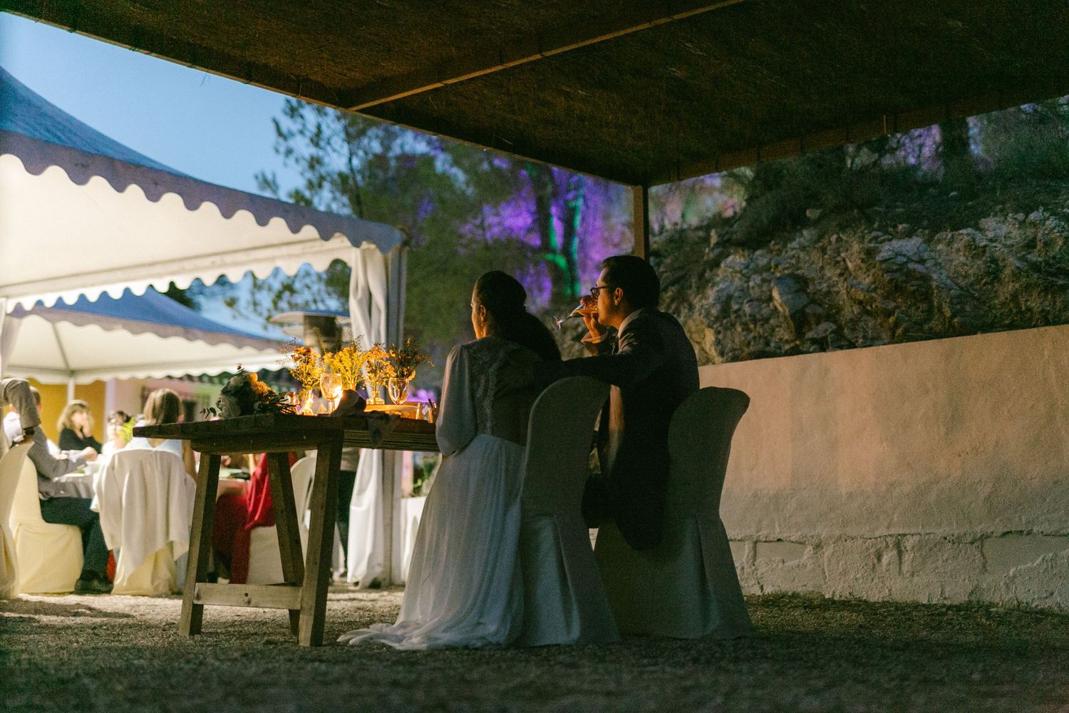 Guests enjoying an outdoor evening reception under a white tent with ambient lighting and stone walls.