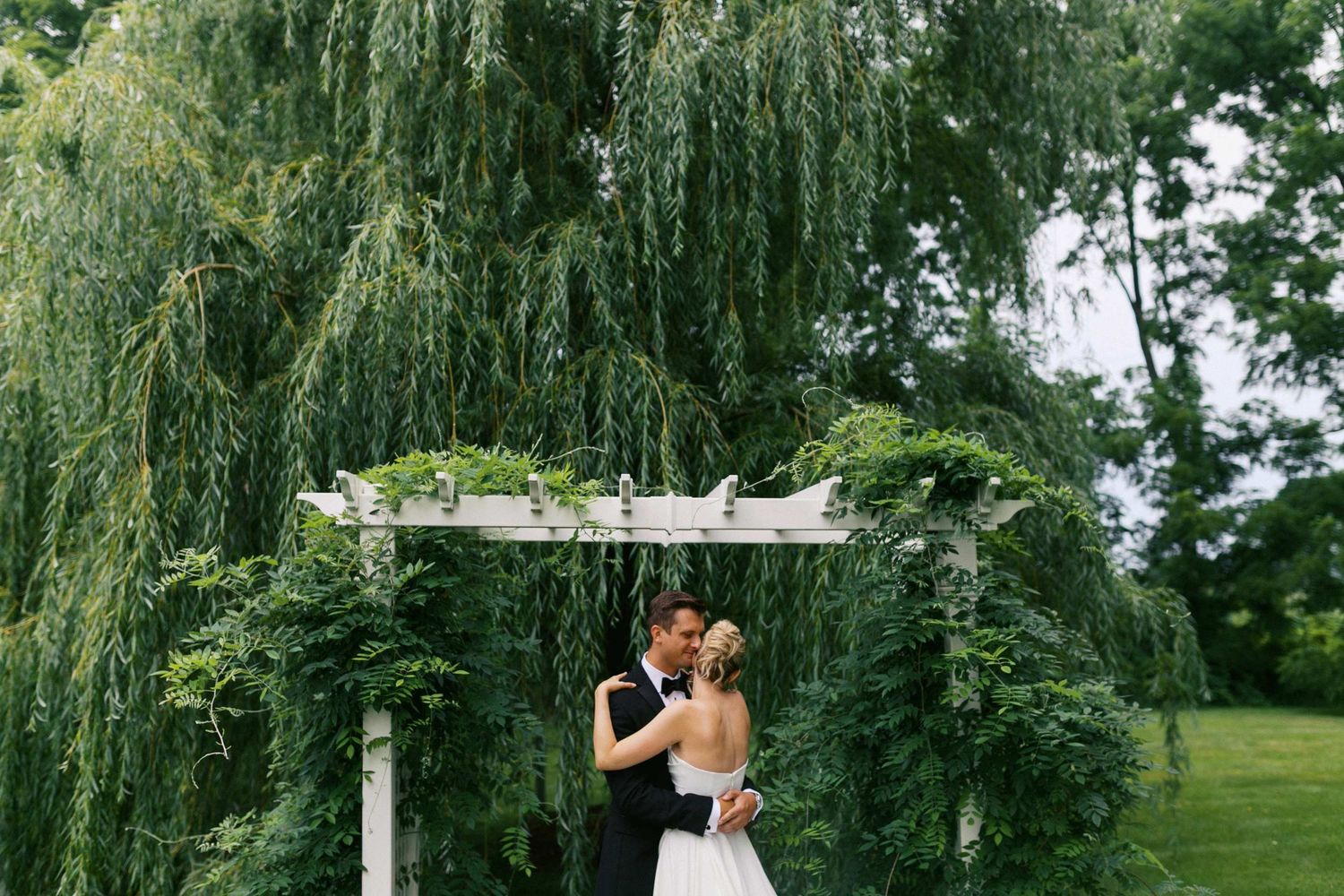 Wedding couple shares romantic moments under weeping willow trees in a lush green garden setting.