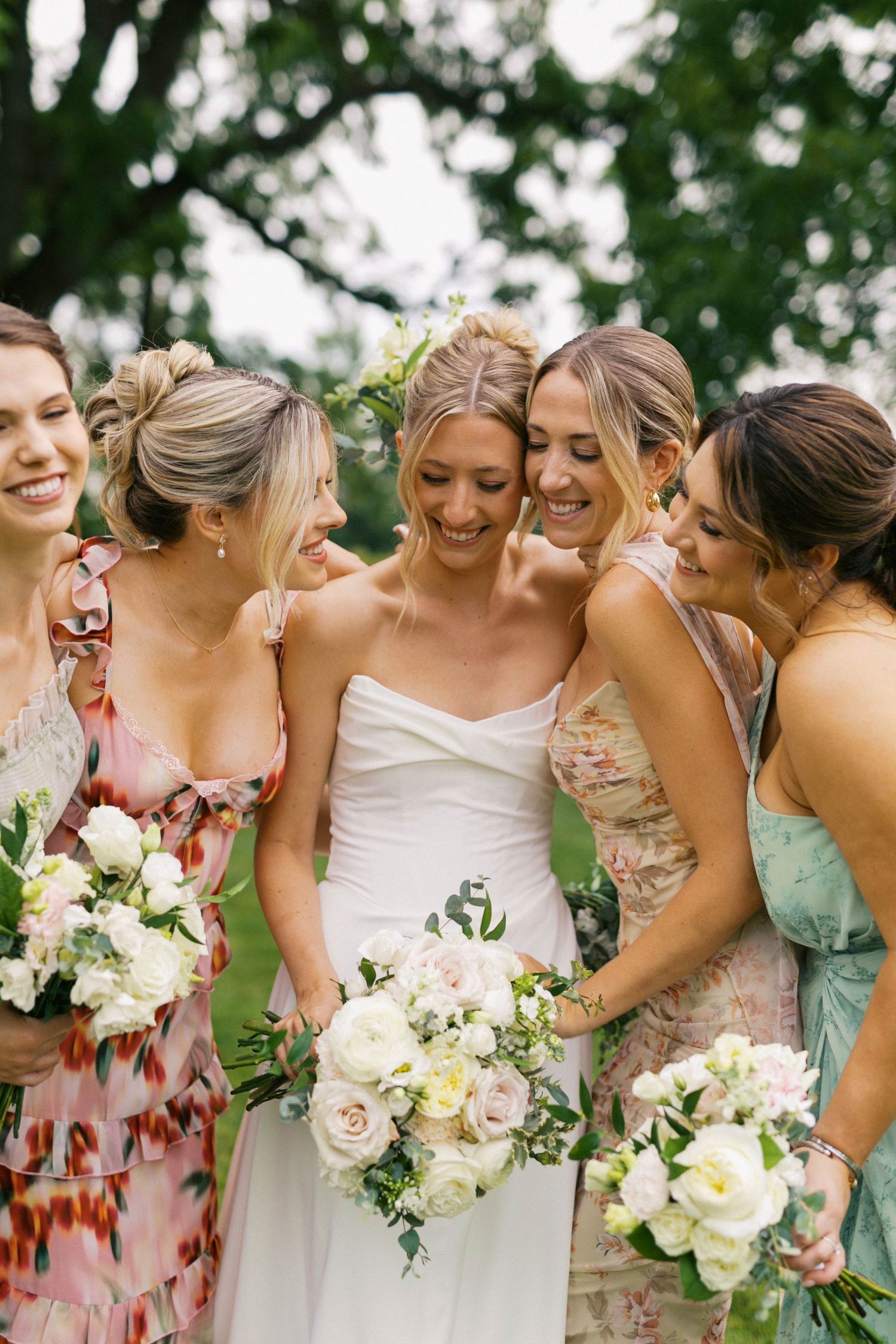A bridal party in pastel floral dresses shares a joyful moment while holding elegant white and blush rose bouquets outdoors.