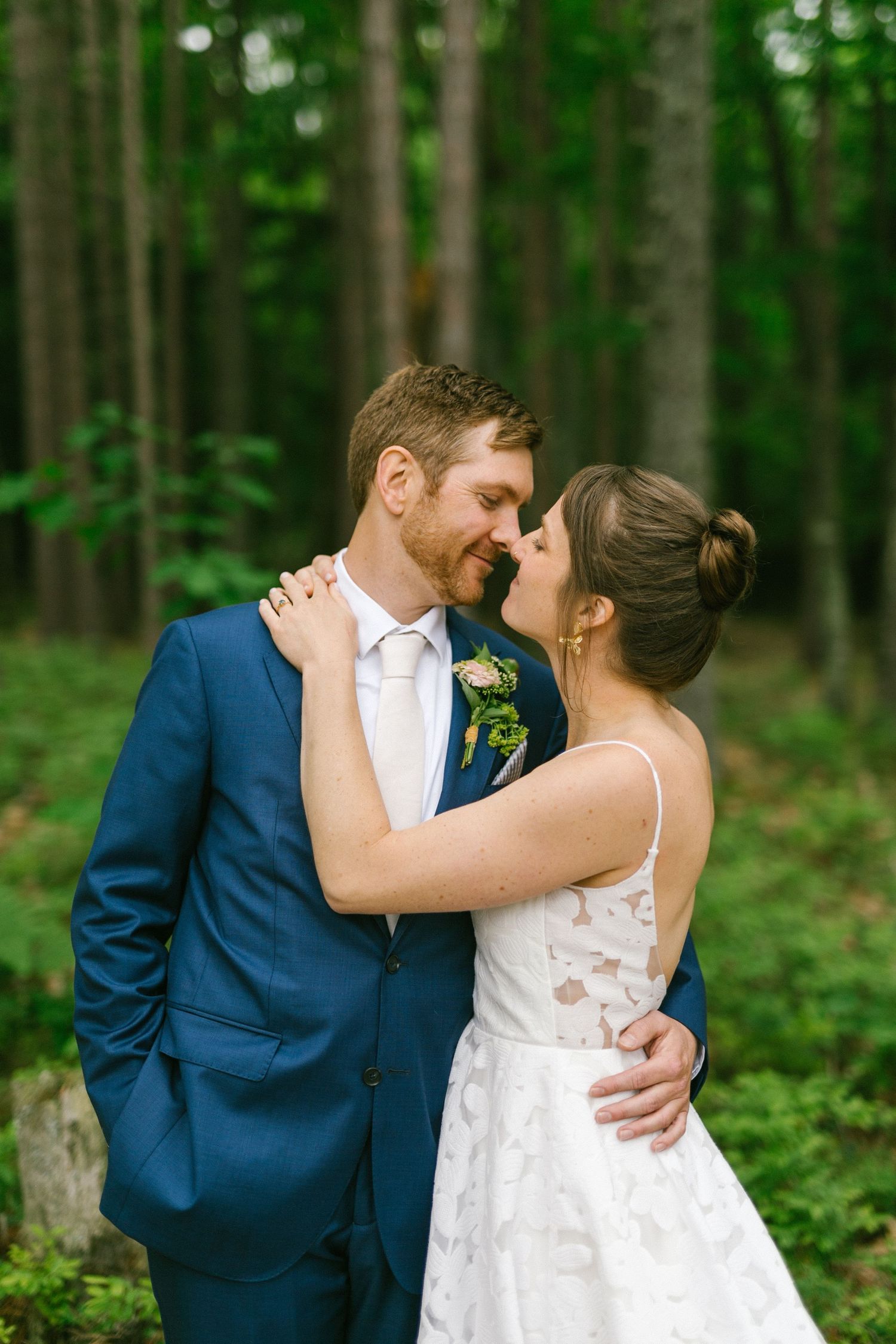 A romantic outdoor wedding moment captured in a lush forest setting with blue suit and white lace dress.