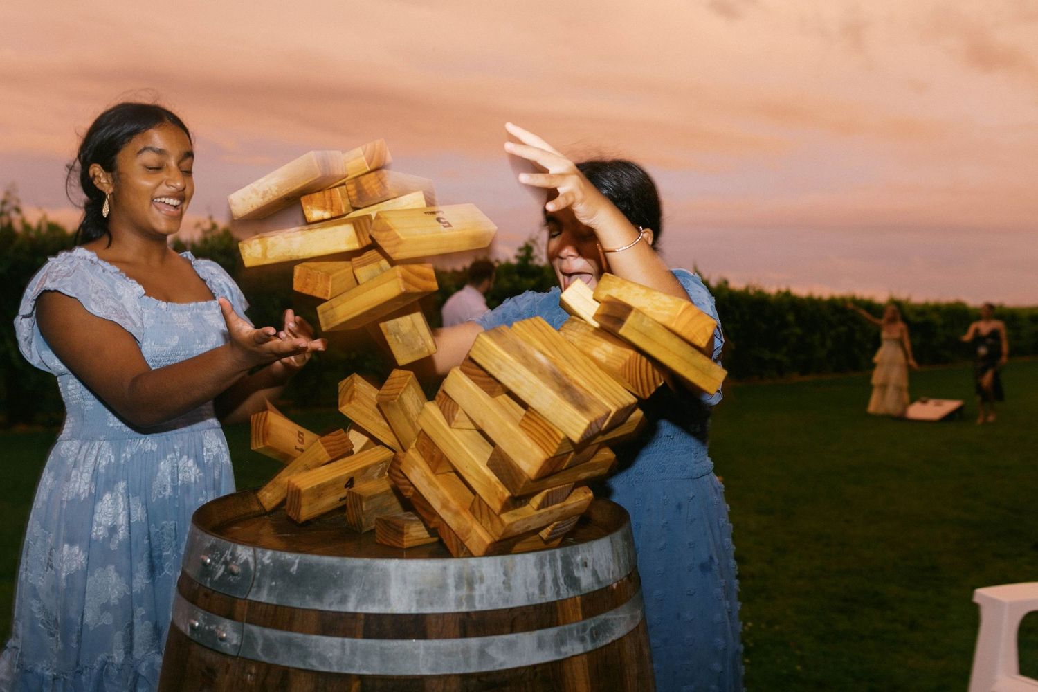 Two young girl cause giant jenga pieces fall onto a rustic wooden barrel at sunset.