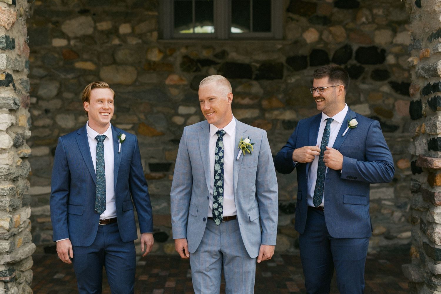 Groomsmen in blue and gray suits gathered outside stone building.