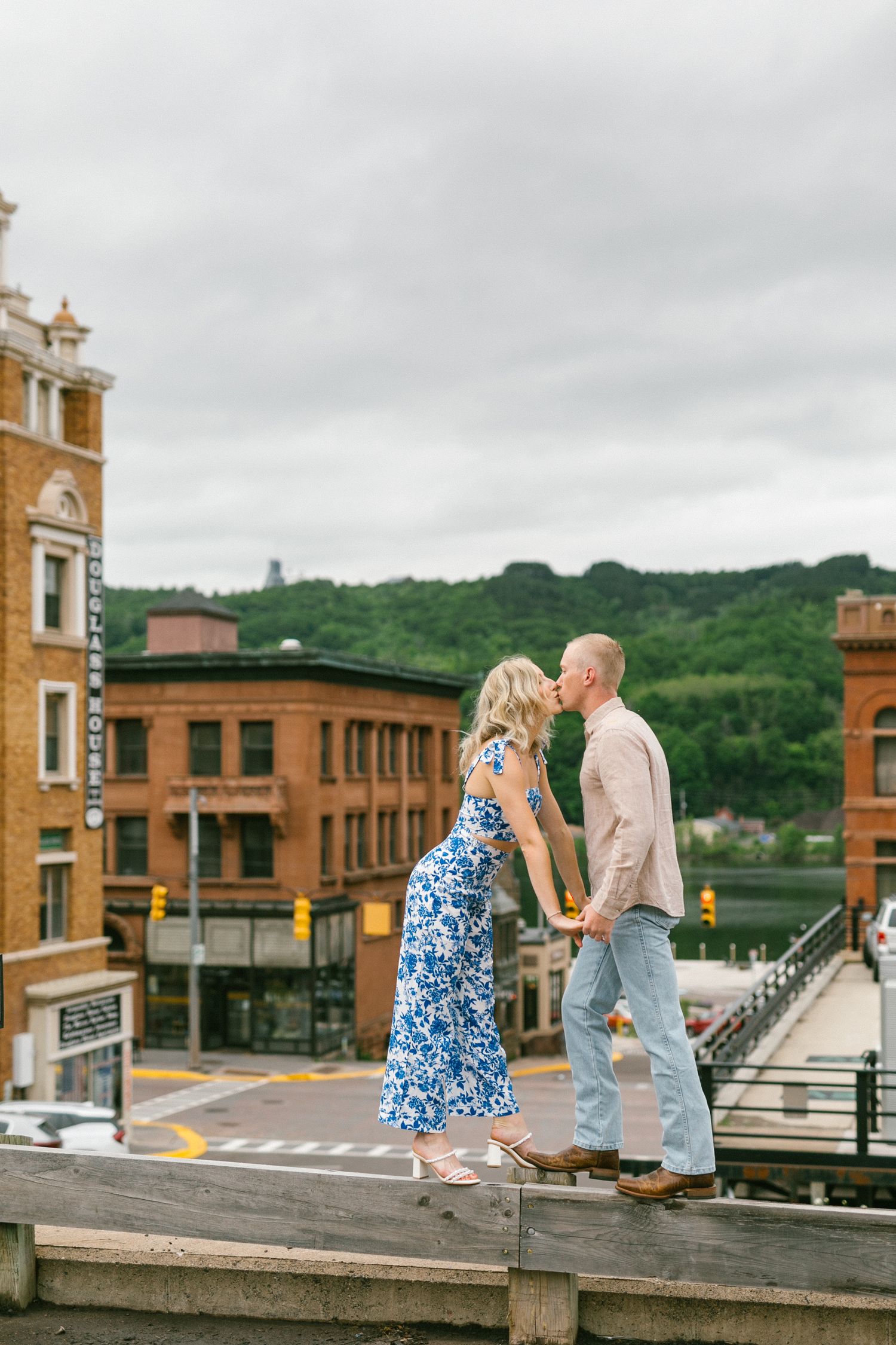 A couple shares a romantic moment on a raised ledge in a historic downtown area with brick buildings and overcast skies.