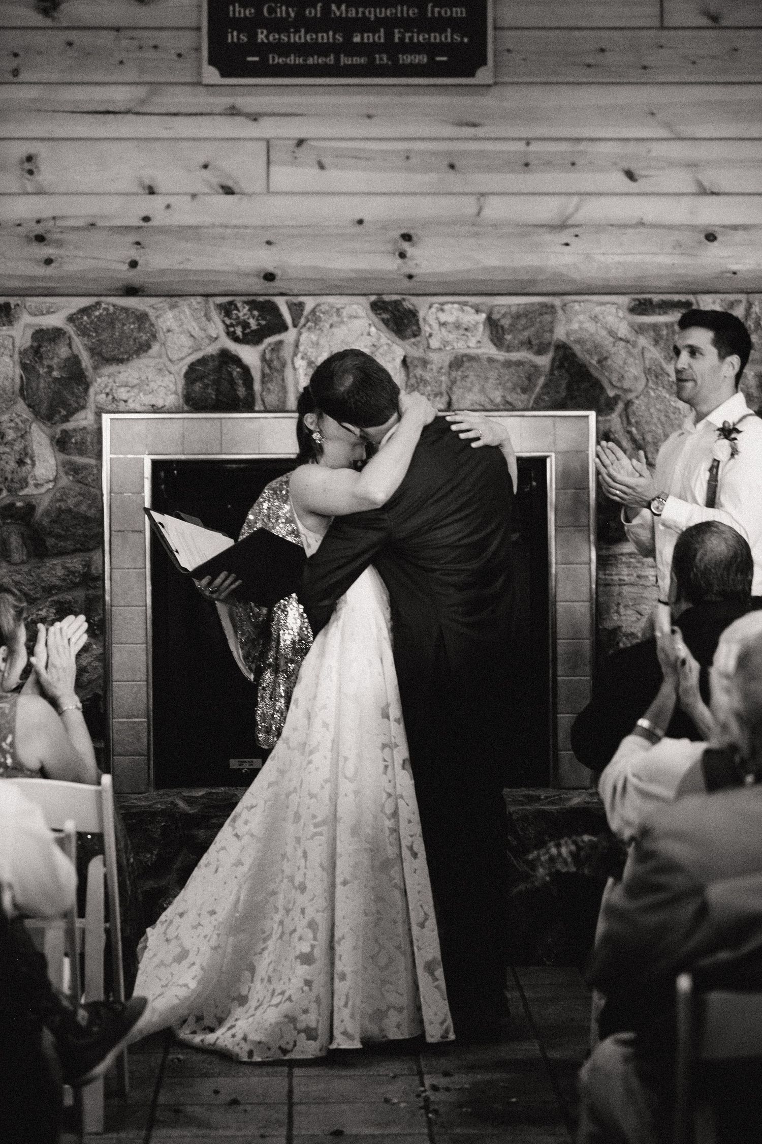 A black and white wedding photo shows a couple embracing in front of a rustic stone wall during an indoor ceremony.