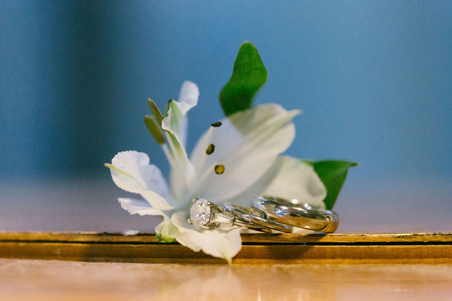 Two gold wedding bands and a diamond engagement ring rest beside a white lily flower on a golden surface.