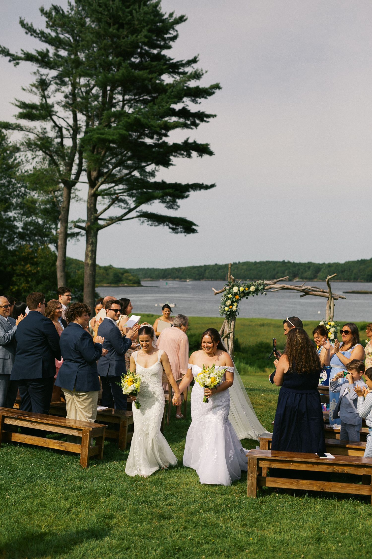 Two brides walk down outdoor aisle holding hands after same-sex wedding ceremony by waterfront.