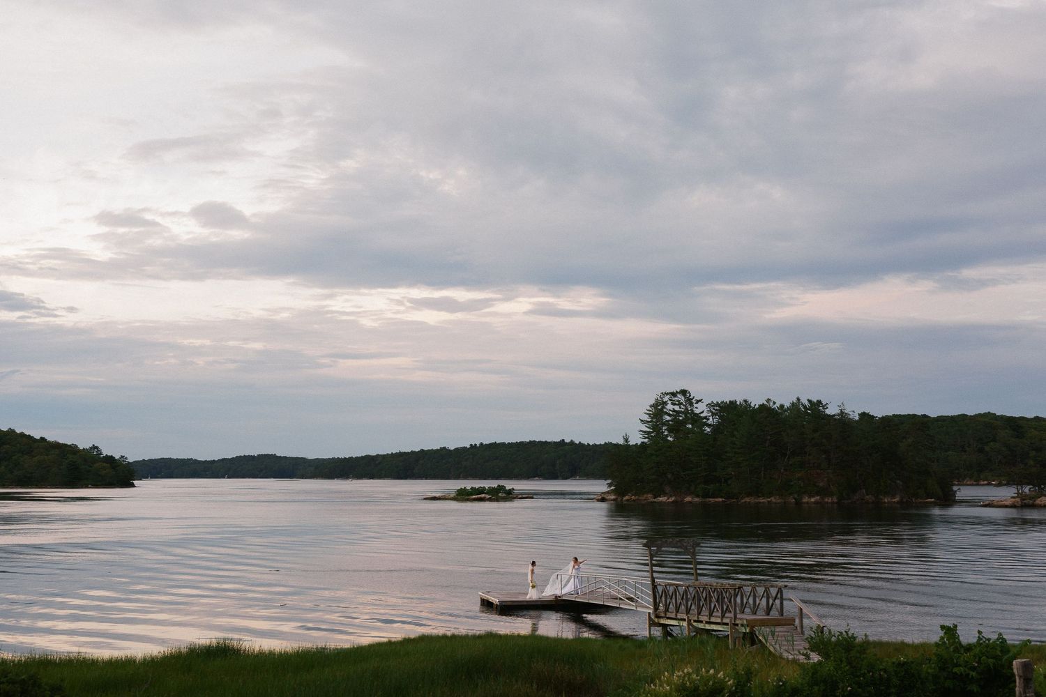 Serene view of the Kennebec River at dusk with a wooden dock, kayaks, lush green islands, and dramatic cloudy sky reflected on calm water.