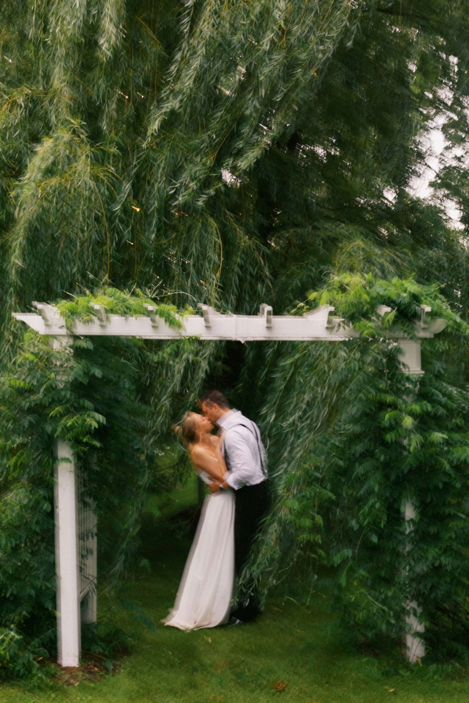 Bride and groom sharing a kiss under a white garden pergola surrounded by lush green weeping willow trees.