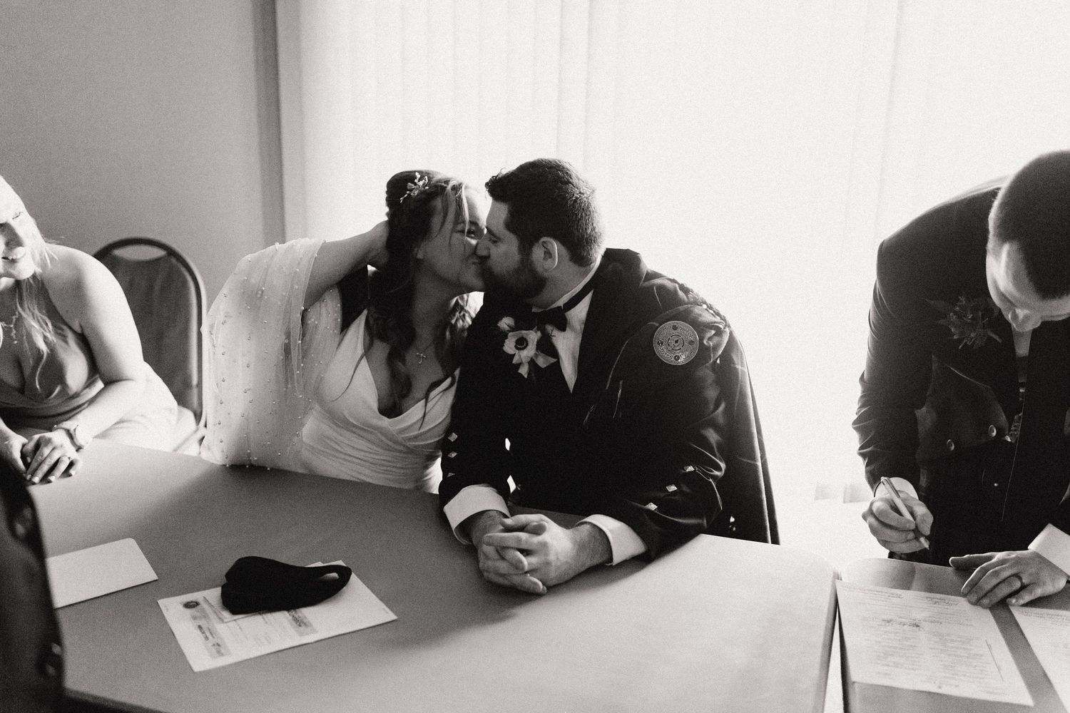 Black and white photo of bride and groom kissing while signing wedding documents at ceremony table.