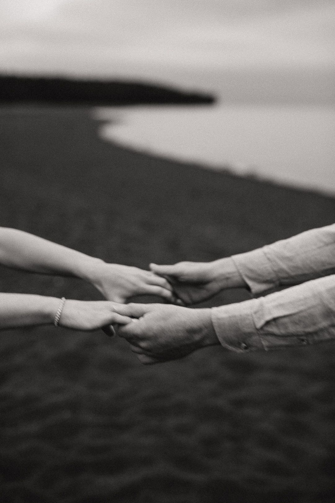 Black and white close-up of couple holding hands on sandy beach shoreline.