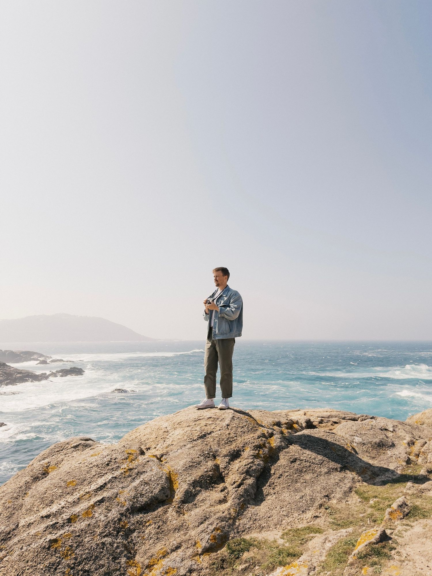 Man standing on coastal rocks overlooking ocean waves under a clear sky.
