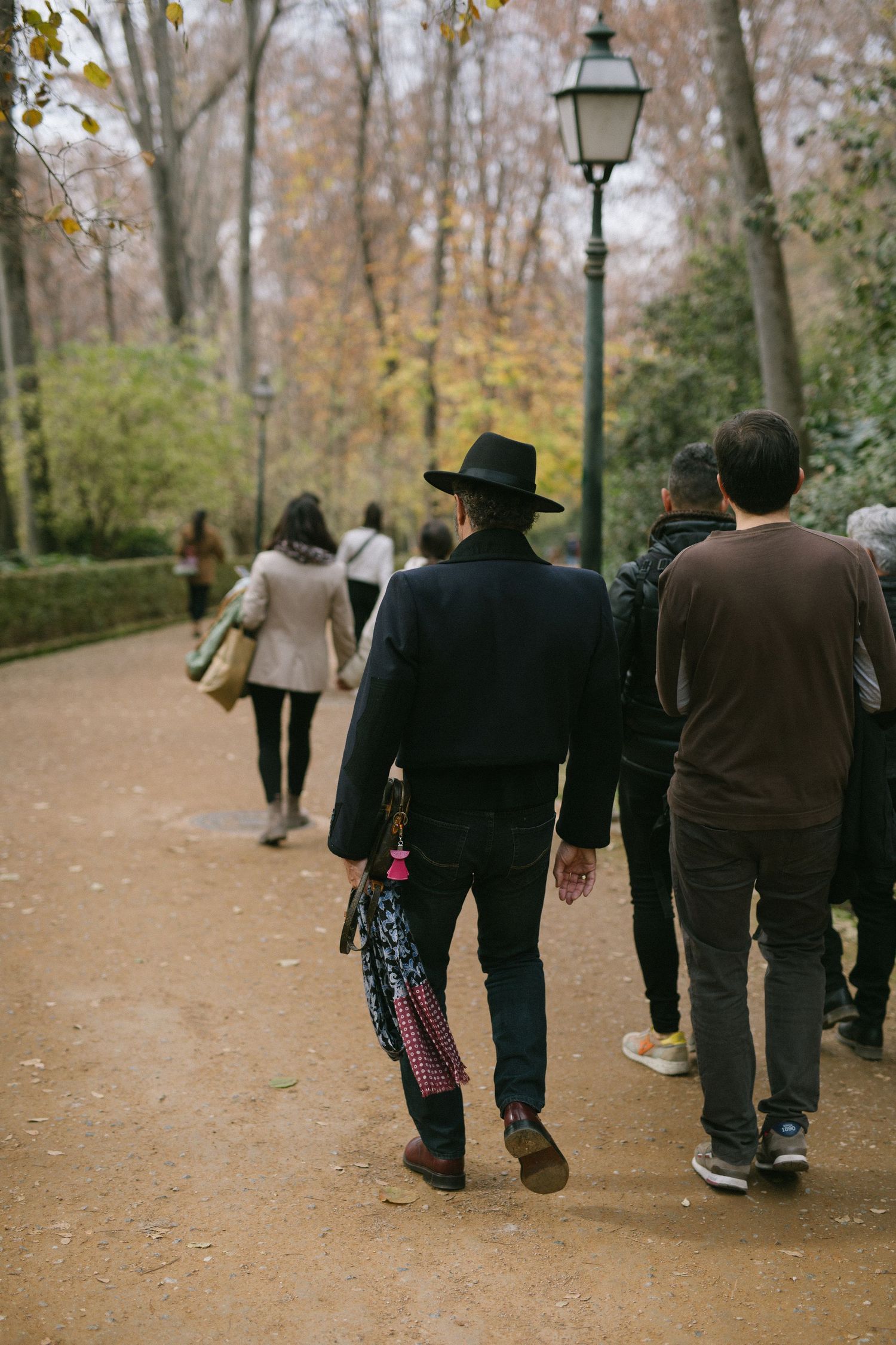 People walking on an autumn park path with fallen leaves and vintage lamp post in Luxembourg Gardens, Paris.