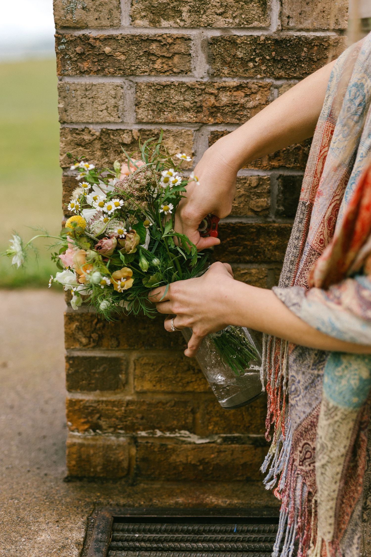 Hands arranging a delicate wildflower bouquet against a rustic brick wall.