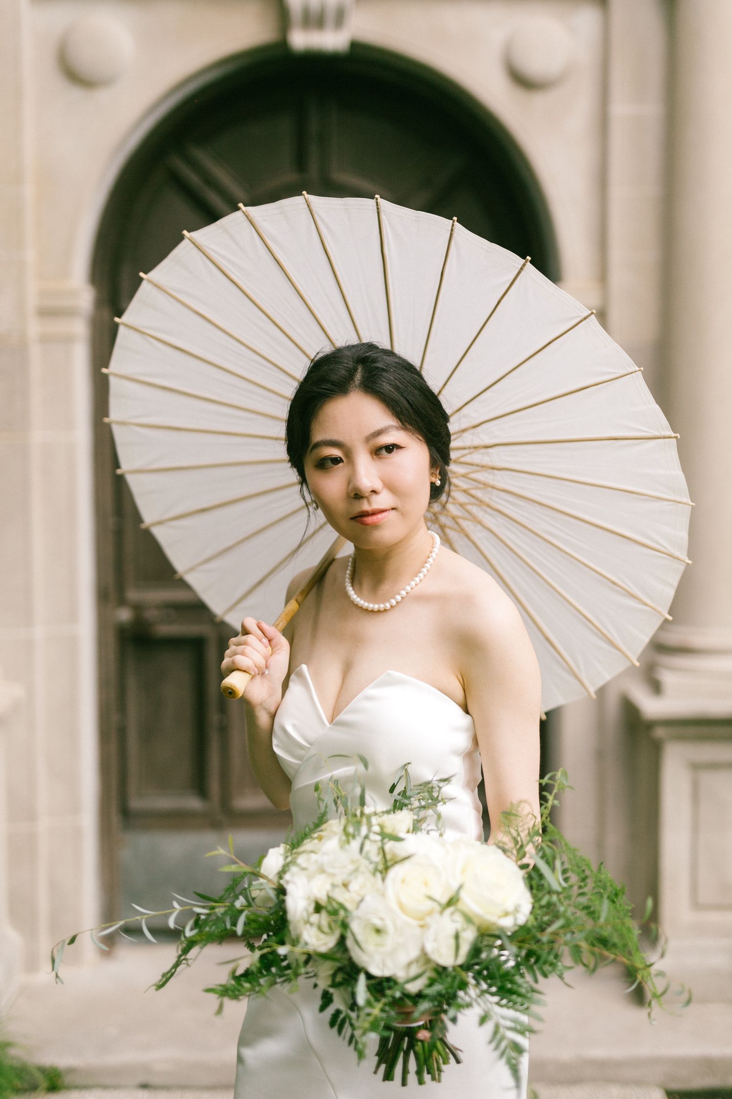 Elegant Asian bride in strapless white gown holding parasol and white floral bouquet outdoors.