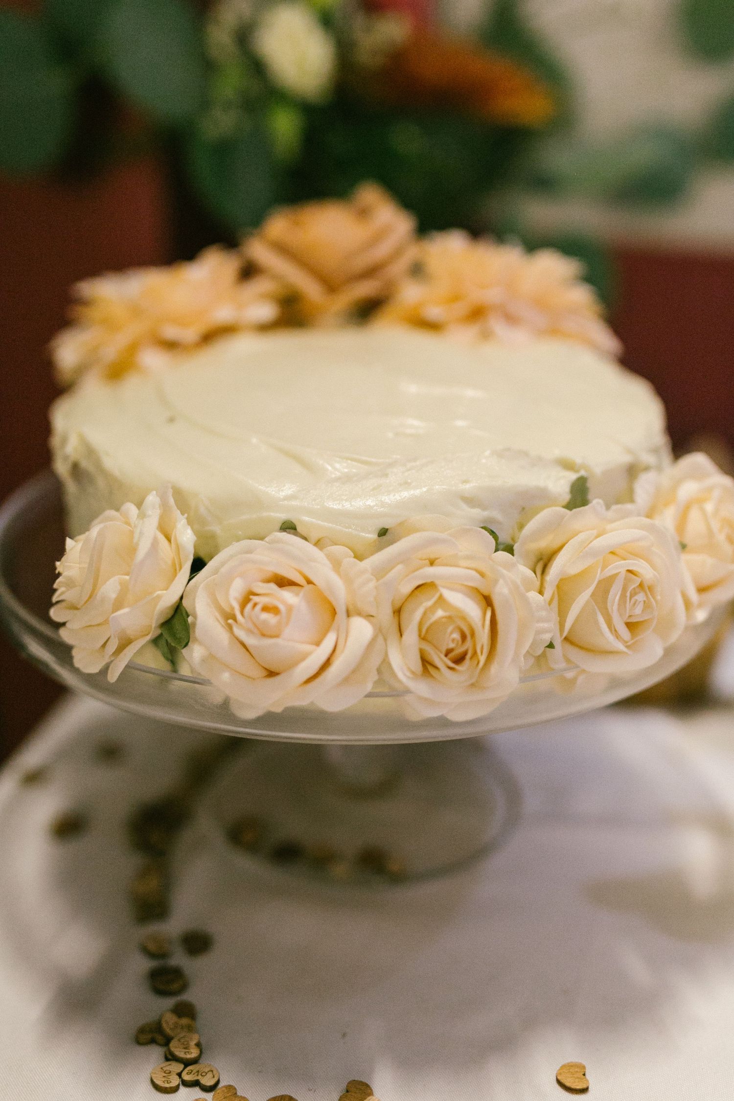 An elegant white frosted cake decorated with pale peach roses sits on a glass pedestal cake stand.
