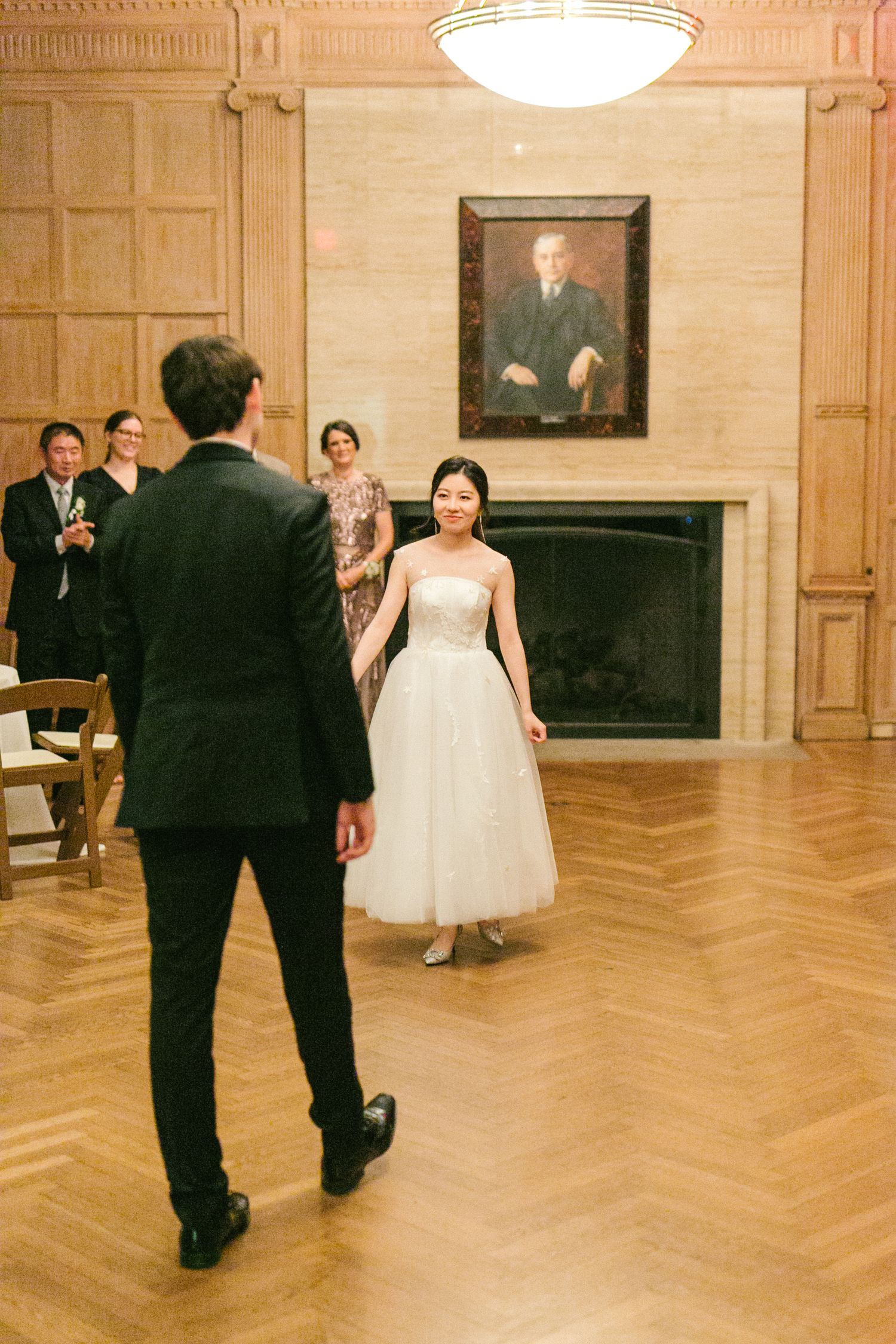 Bride in white tulle dress walks toward groom in black suit during first dance in elegant wood-paneled ballroom.