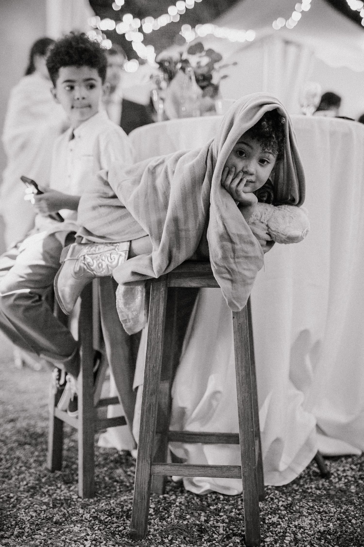 Black and white photo of children resting on wooden stools at an event with string lights and white decor in background.