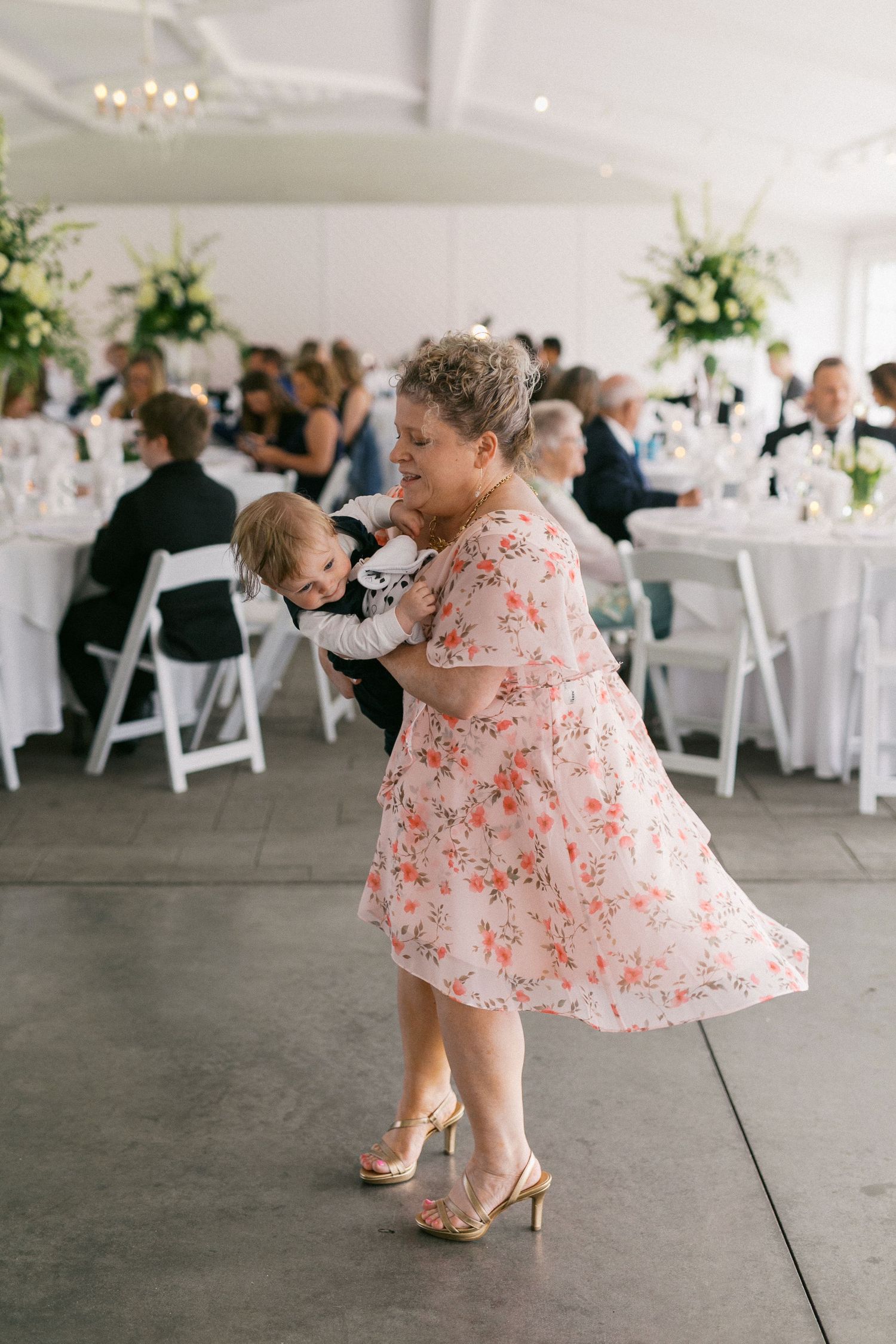 A woman in a floral dress dances while holding a baby at a wedding reception.